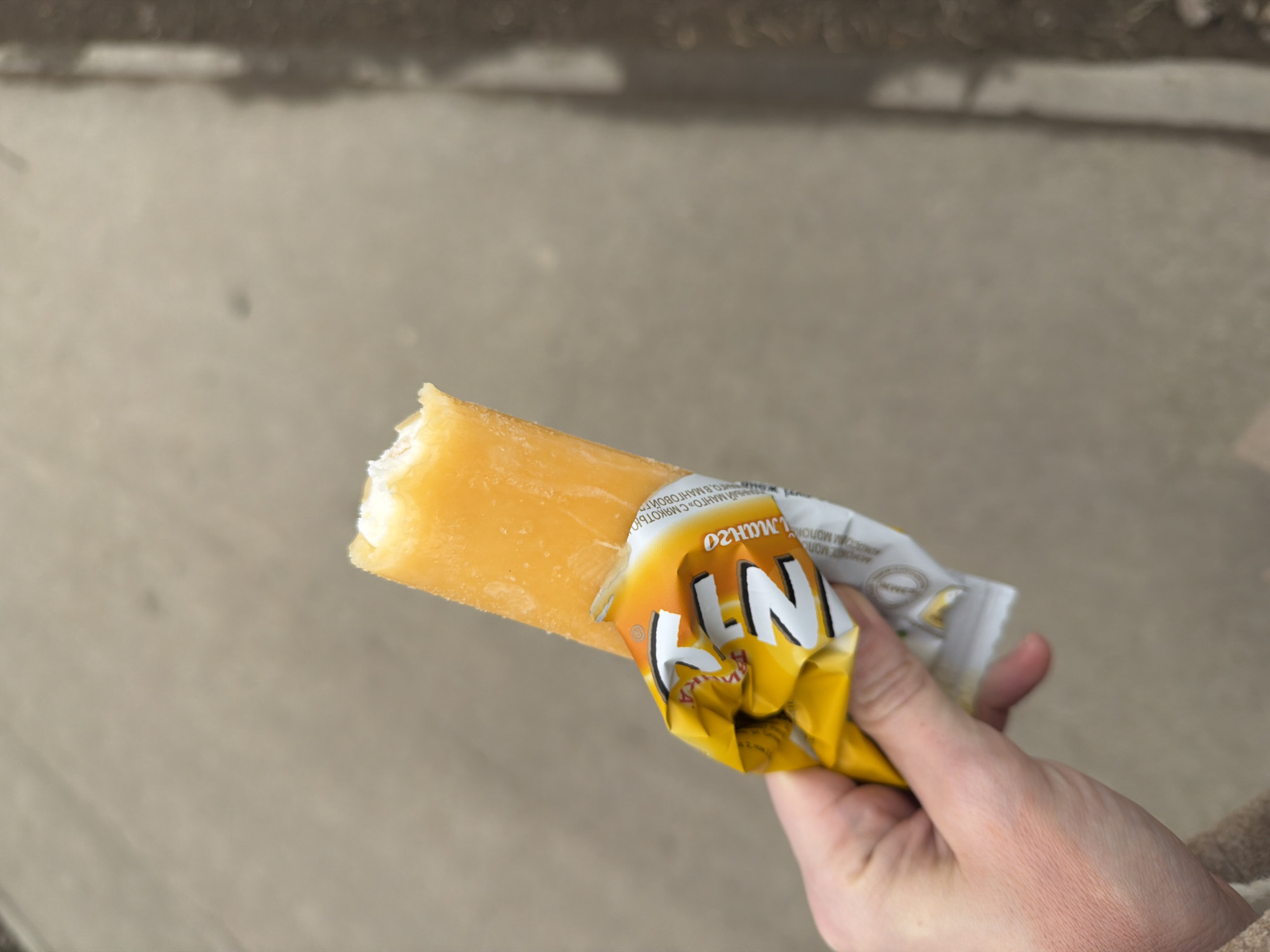 The image captures a close-up, high-angle view of a pale-skinned human hand holding a partially consumed orange-colored frozen confection, identified as a mango-flavored ice cream bar by the Russian text "МАНГО" (MANGO) visible on its crumpled yellow and white wrapper. The top portion of the bar displays a bite mark, revealing a lighter, possibly white, interior. The background consists primarily of light gray pavement or asphalt. On the far right, a dark, blurred vertical element and a narrow strip of darker ground material are visible. The scene is located in Perm, Russia.