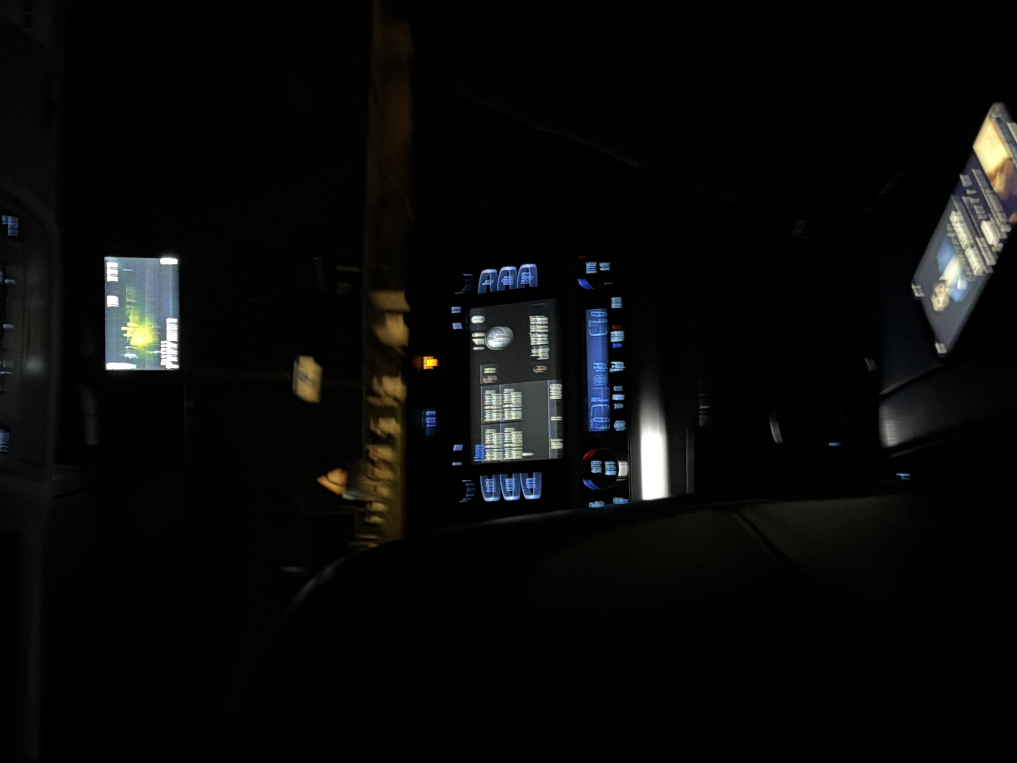 The image shows the interior of a vehicle at night. The infotainment system is visible on the dashboard, displaying various information. In the back, a passenger entertainment screen is visible, angled towards the viewer. Outside the window is the blur of the urban environment at night, with lights of buildings visible. The foreground is dominated by the rear seat of the car. It is impossible to verify the image's location is in Kuchak, Armenia.