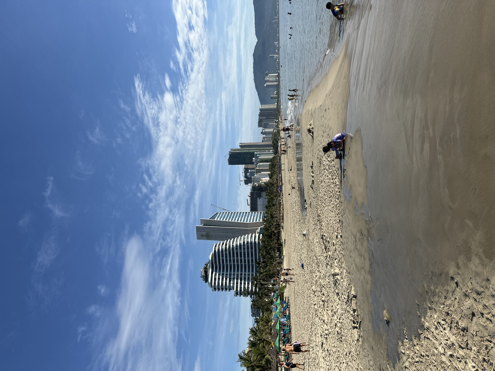 This is a wide-angle shot of a busy beach in Đà Nẵng, Vietnam, with the ocean on the right and a row of modern high-rise buildings in the background, including the distinctive curved "Dragon Bridge" hotel. The sand is light brown and scattered with footprints. There are several people on the beach: a group of children are playing near the water's edge, digging in the wet sand, while other individuals are walking along the shore or bathing in the sea. In the mid-ground, lounge chairs and umbrellas suggest a resort area. Palm trees line the back of the beach, adding to the tropical atmosphere. The sky is a vibrant blue with scattered white clouds, indicating fair weather. The overall scene depicts a lively beach day in a developed coastal city.