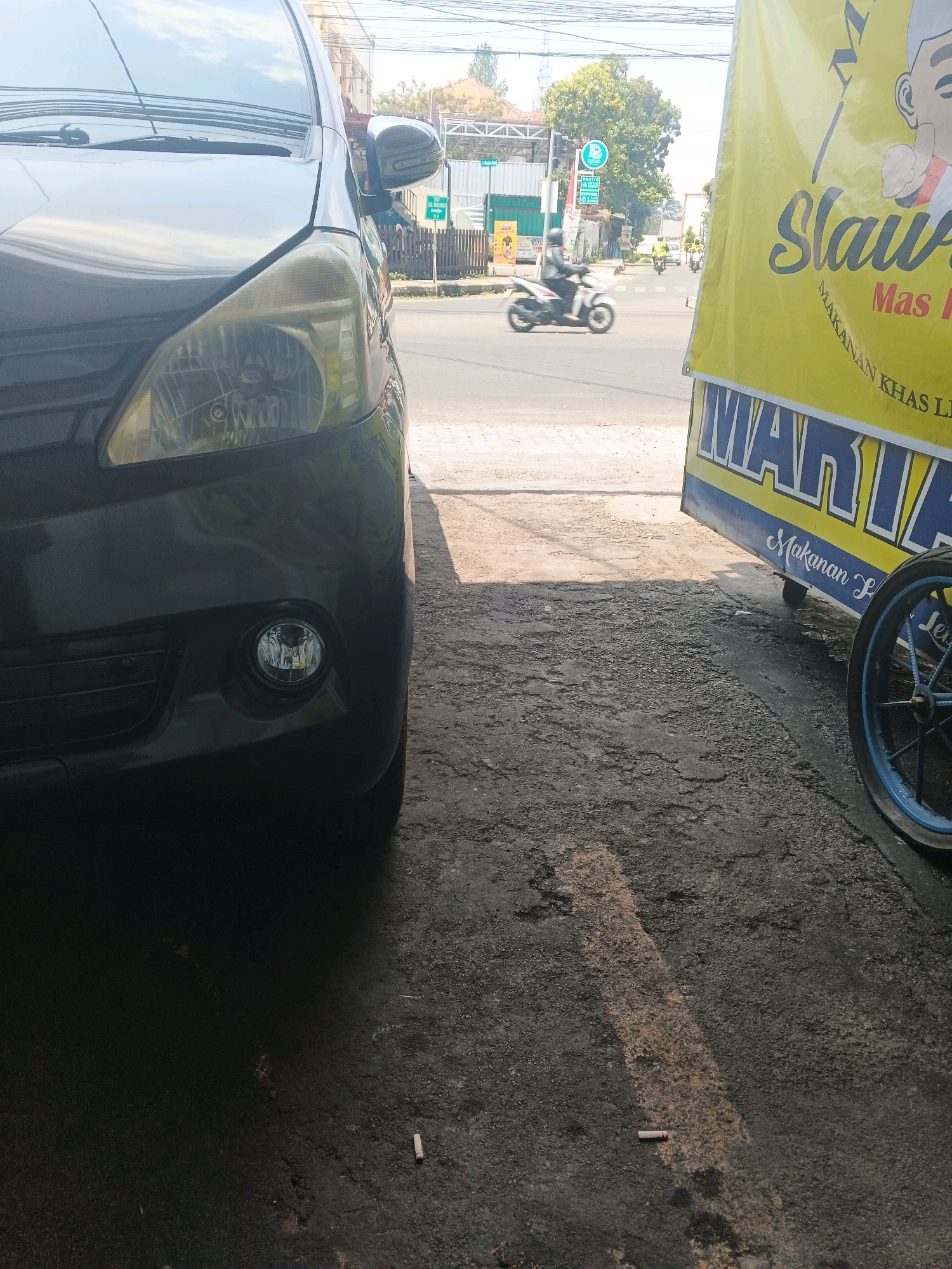 In Bandarjo, Indonesia, a black car is parked near a food cart. The car's headlight and fog light are visible on the left side of the frame. The food cart, with a yellow banner advertising "Slawut Mas L" and "MAKANAN KHAS LI," is on the right. The cart has a blue wheel with spokes. The ground is asphalt, with a faint shadow cast from the car. In the background, a street scene unfolds with a traffic light, trees, and several motorcycles in motion. One motorcycle is in the foreground, moving from left to right. Power lines are visible against the sky.
