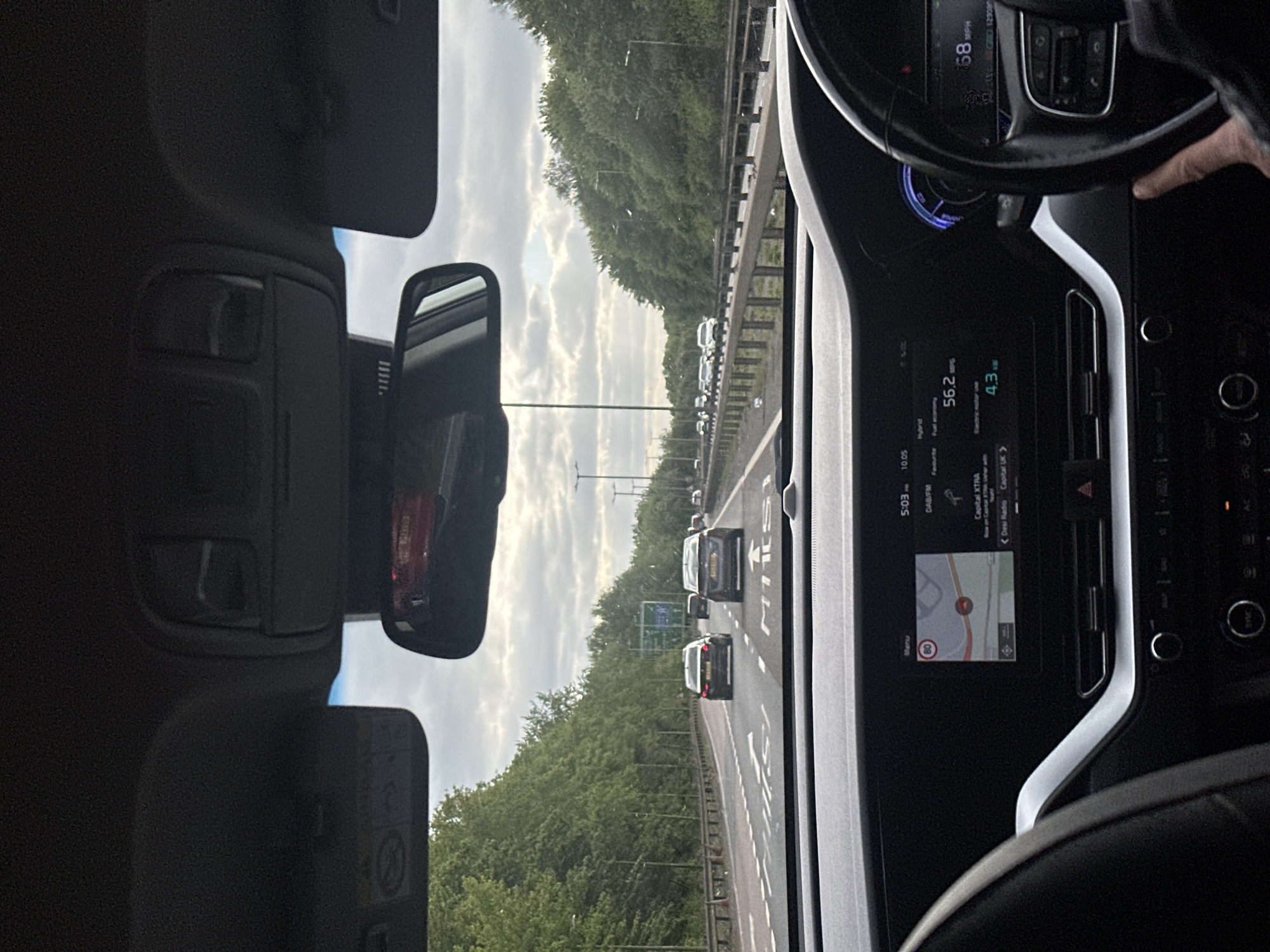 The image depicts a car interior, specifically the driver's perspective. The car is on a highway, with other vehicles visible ahead. The dashboard displays information such as speed (68 mph), fuel economy (56.2 mpg), and a navigation map indicating a speed limit of 80. The radio is tuned to Capital XTRA. The environment appears to be a wooded area alongside the highway, under a cloudy sky. The location is likely within Uttlesford, United Kingdom, based on the "Capital UK" radio station selection. A person's hand is visible on the steering wheel.