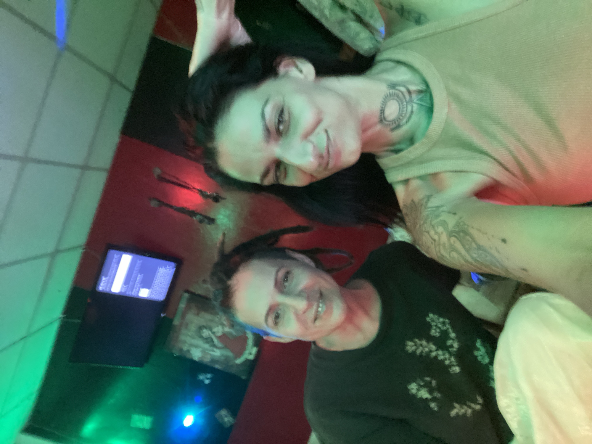 Two adult females pose for a close-up selfie photograph inside a dimly lit establishment, likely a bar or lounge, in Mũi Né, Vietnam. The scene is illuminated by strong colored lighting, primarily green from the ceiling and a blue spotlight on the left, casting a red hue on the rear wall.
The woman on the left, positioned slightly behind and to the left of the other, has light skin, dark hair styled into dreadlocks pulled back from her face, and is smiling directly at the camera, revealing a slight gap in her upper front teeth. She wears a dark, long-sleeved shirt with light-colored floral or botanical patterns.
The woman on the right, closer to the camera and presumably holding the device for the selfie, also has light skin, long dark hair, and a slight smirk while looking towards the camera. She has visible tattoos, including an intricate circular design on her neck and additional patterns on her left shoulder and arm. She wears a light-colored (possibly tan or beige) ribbed tank top. Her left hand is partially visible near her head, resting on her hair.
The background features a red-painted wall with a darker section to the left. Mounted on the walls are two flat-screen monitors; the left one is dark, while the right one, mounted vertically on the red wall, displays white text on a blue or purple interface. Below the monitors, a framed picture or artwork depicting a figure in a red dress is visible. Further right on the red wall, two elongated, dark, decorative objects are mounted. The ceiling consists of white grid tiles, heavily washed in green light. A bright blue light source is visible on the far left.