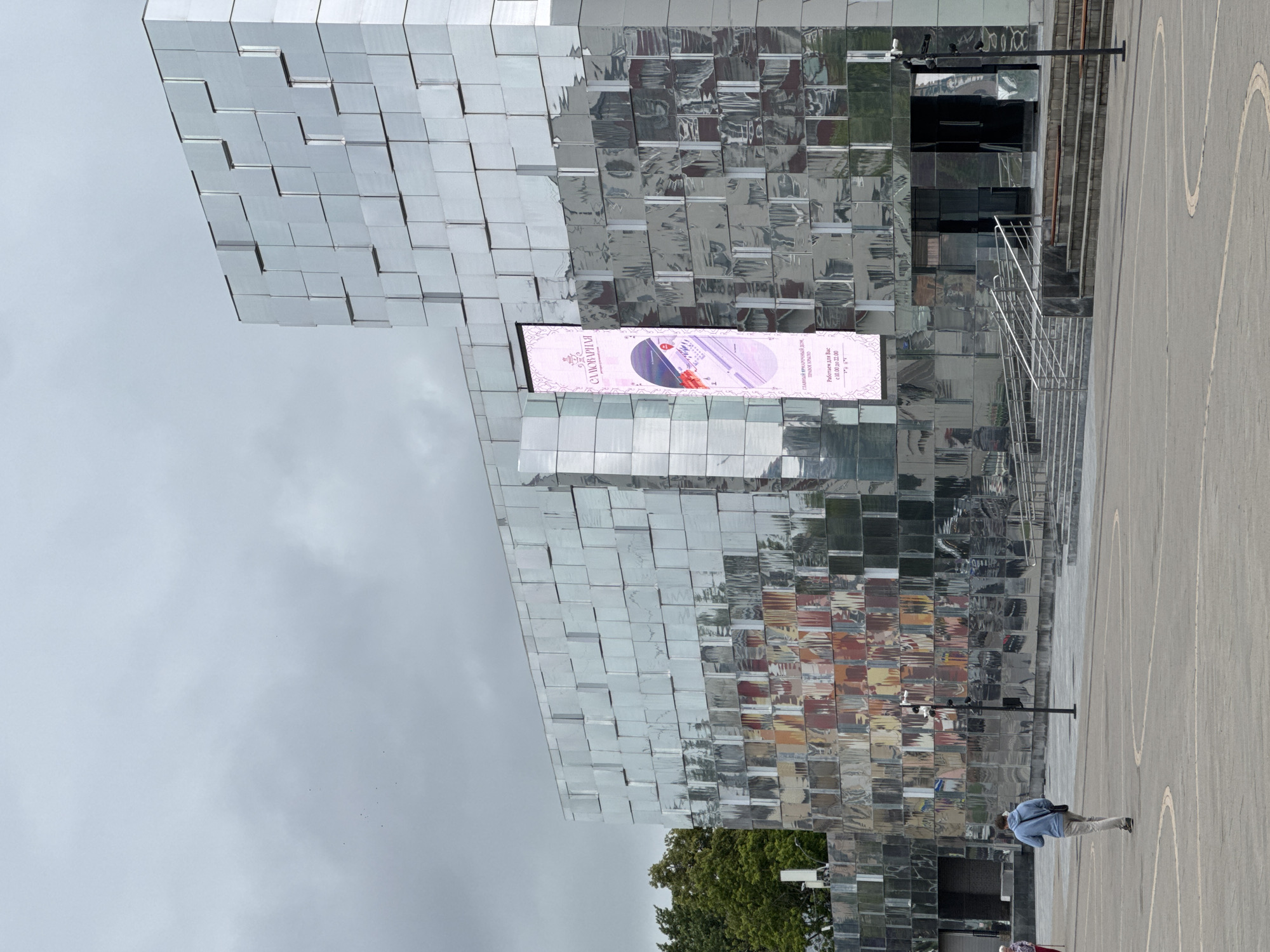 The image depicts the exterior of the "Samovar House" located in Nizhny Novgorod, Russia. The building features a distinctive facade composed of numerous reflective, silver-colored rectangular panels, giving it a mosaic-like appearance. A large vertical digital banner is displayed on the building's surface, showing an advertisement with text in Russian.
In the foreground, a paved open area is visible, with a person walking away from the camera, dressed in a blue hoodie and light-colored pants. The person is carrying a bag over their shoulder. To the left of the frame, a group of trees can be seen. The environment appears to be an urban public space, possibly a plaza or square, with the building forming the backdrop. The sky is overcast. The digital banner on the building shows an illustration of a woman in a hat and includes text that appears to be an address and operating hours. Stairs and railings leading to an entrance are also visible on the right side of the building.