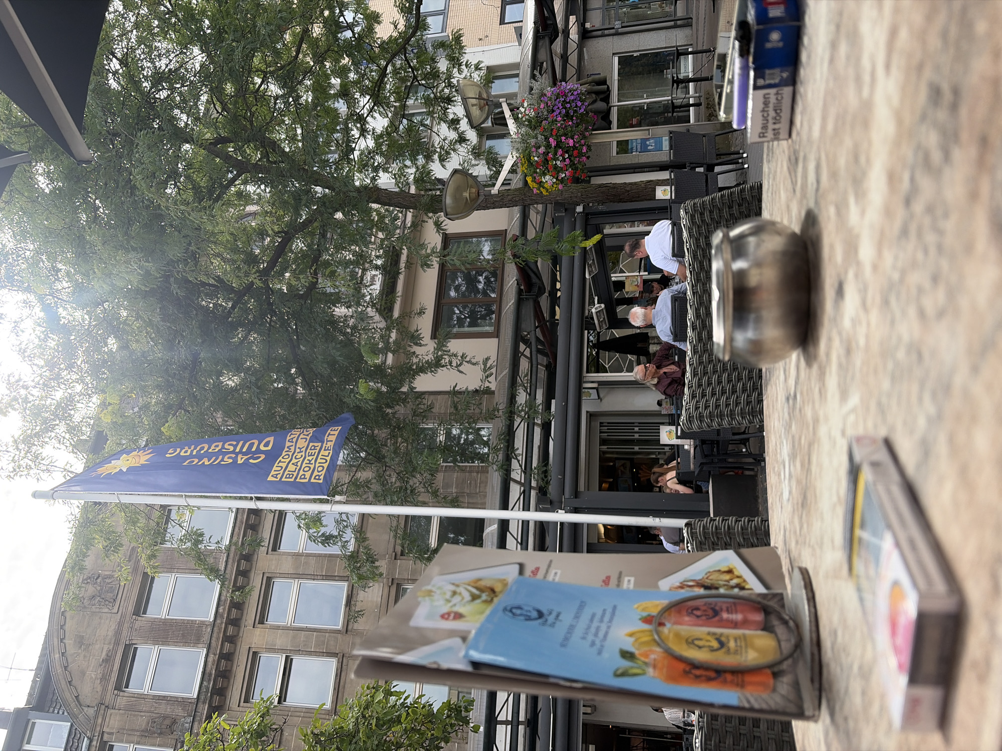 The image depicts an outdoor seating area of a casino in Duisburg, Germany, with a blue flag prominently displayed, bearing the text "CASINO DUISBURG" along with "AUTOMATIC BLACK JACK POKER ROULETTE". The foreground shows a table with a pack of cigarettes and an ashtray, suggesting a smoking-friendly environment. In the background, patrons are seated at tables, some engaged in conversation, while others appear to be dining or observing the surroundings. The architecture of the buildings in the background indicates a European urban setting. A lush green tree partially obscures the view of the casino's facade. The overall atmosphere suggests a casual daytime gathering at an establishment that offers gaming and refreshments.