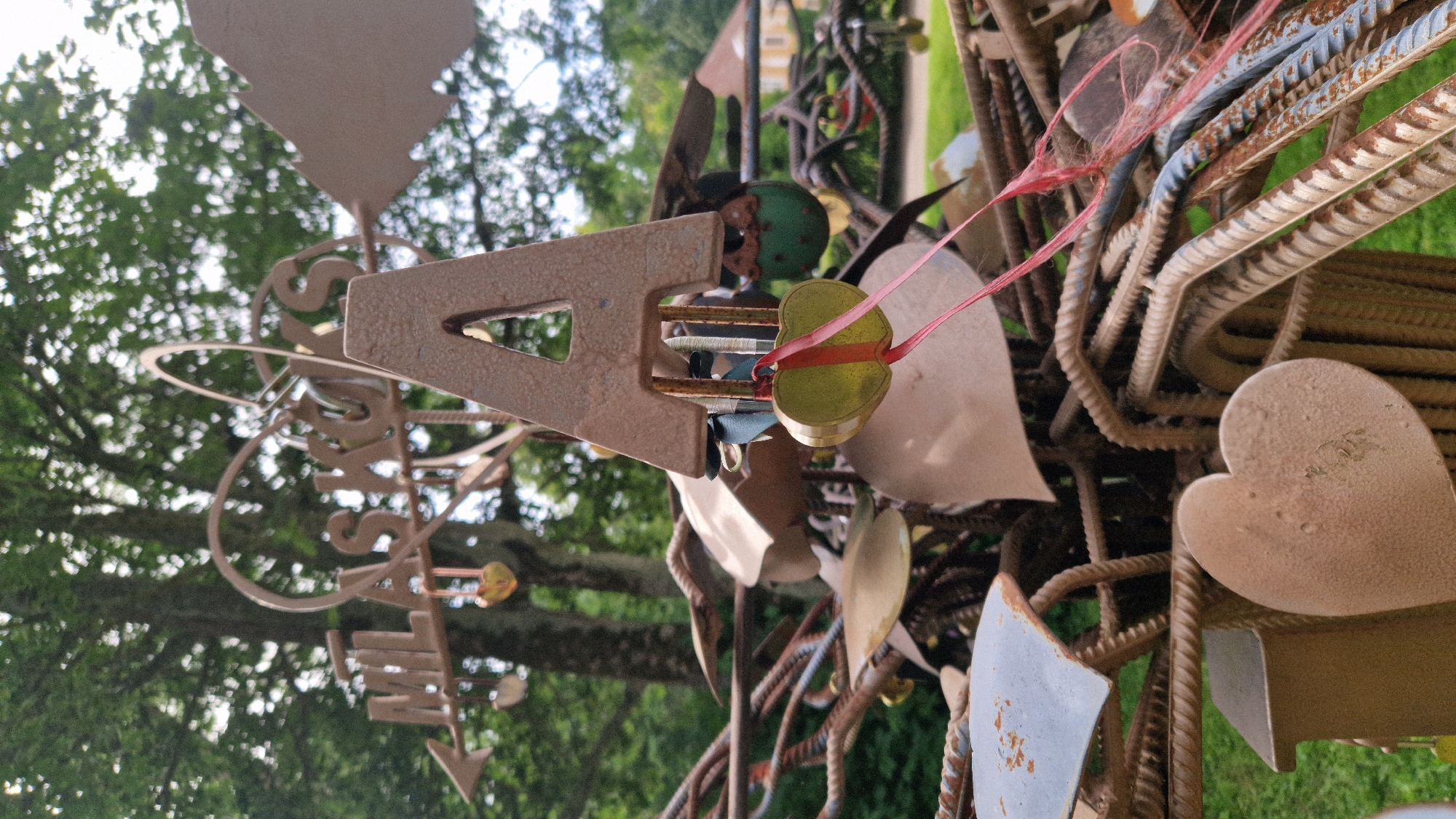 The image shows a close-up of a metal sculpture adorned with heart-shaped locks, likely a lover's lock sculpture, situated in a park or outdoor setting. The sculpture features an array of metal elements, including what appears to be a stylized wind vane with the letters "MILAS KOKS" visible, a large letter "A", and numerous heart-shaped locks of various sizes and colors, some hanging by ribbons. The background consists of lush green trees and foliage, suggesting a natural environment. In the distance, a building with yellow siding and a red roof is partially visible. The sculpture appears to be made of rebar and other metal pieces, giving it a rustic and somewhat industrial aesthetic. The focus is on the intricate details of the sculpture and the locks. The scene is well-lit, indicating it was taken during daylight.
