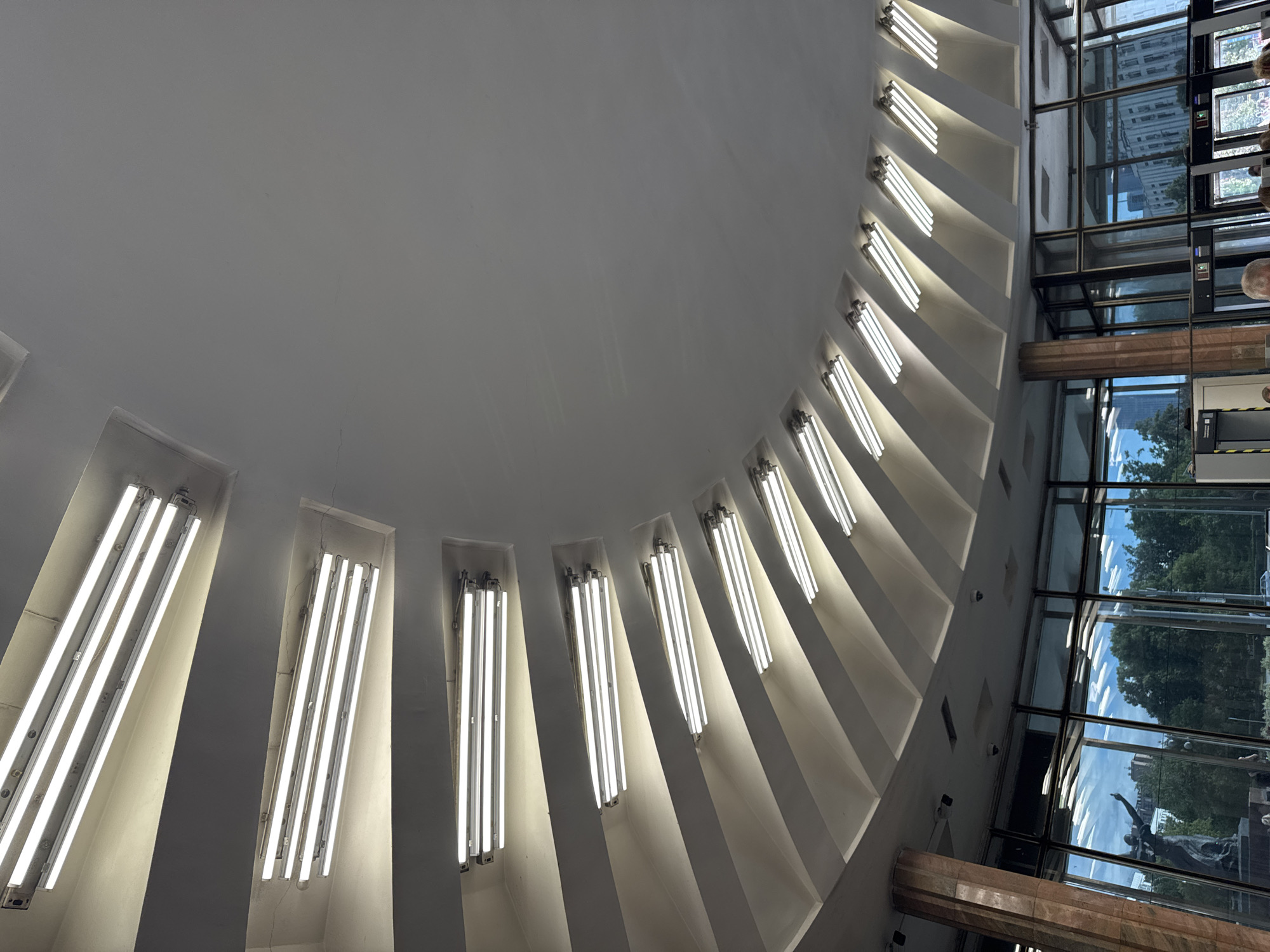 The image shows an interior view of a large, modern building with a distinctive circular ceiling design. Numerous fluorescent tube lights are embedded in angled ceiling recesses, creating a radiating pattern. The ceiling itself is a smooth, white surface. Below the ceiling, large glass windows offer a view of the exterior, which includes trees, buildings, and a statue. A large, marble-clad column is visible on the left side of the frame, supporting the ceiling structure. In the foreground, parts of the interior can be seen, including what appears to be a security checkpoint with metal detectors and people walking through. The overall impression is that of a public building, possibly a museum, exhibition hall, or transport hub, characterized by its grand, illuminated architecture. The scene is located in Moscow, Russia.