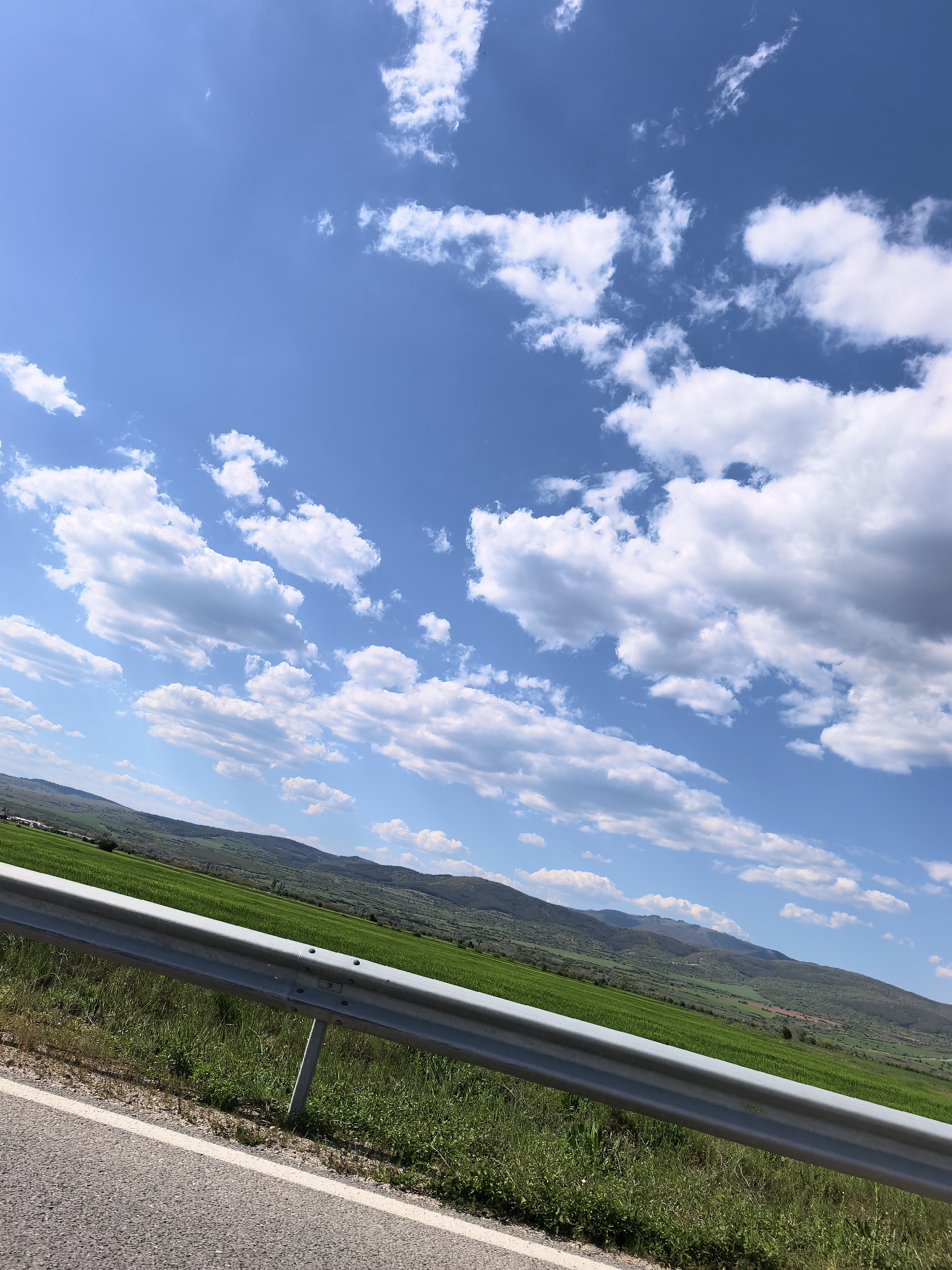 The image shows a landscape with a road in the foreground and a field of green hills in the background under a blue sky with scattered white clouds. The road is made of asphalt with a white painted edge line. A silver guardrail runs parallel to the road. Beyond the guardrail is a field of vibrant green vegetation, leading to gently sloping hills in the distance. The hills are covered with a mix of greenery and brown patches. The sky is blue with puffy white clouds scattered throughout. The scene is likely taken from a vehicle, and based on the information provided, it is located in or near Yahinovo, Bulgaria. There are no people, apparent actions, or interactions captured in the frame.