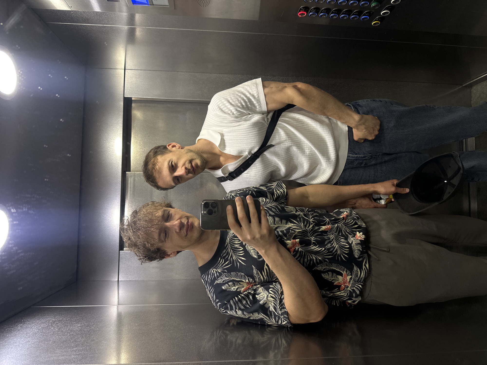 Two men stand in an elevator, taking a selfie. The man on the left, with curly blonde hair, wears a dark t-shirt with a floral pattern. He holds a smartphone in his right hand, angled to capture the reflection. He also has a can in his left hand and a black hat on the brim. The man on the right, with short brown hair, wears a white ribbed polo shirt and blue jeans. A black strap from a bag is slung across his chest. The elevator has stainless steel walls and a panel of illuminated buttons on the right side.