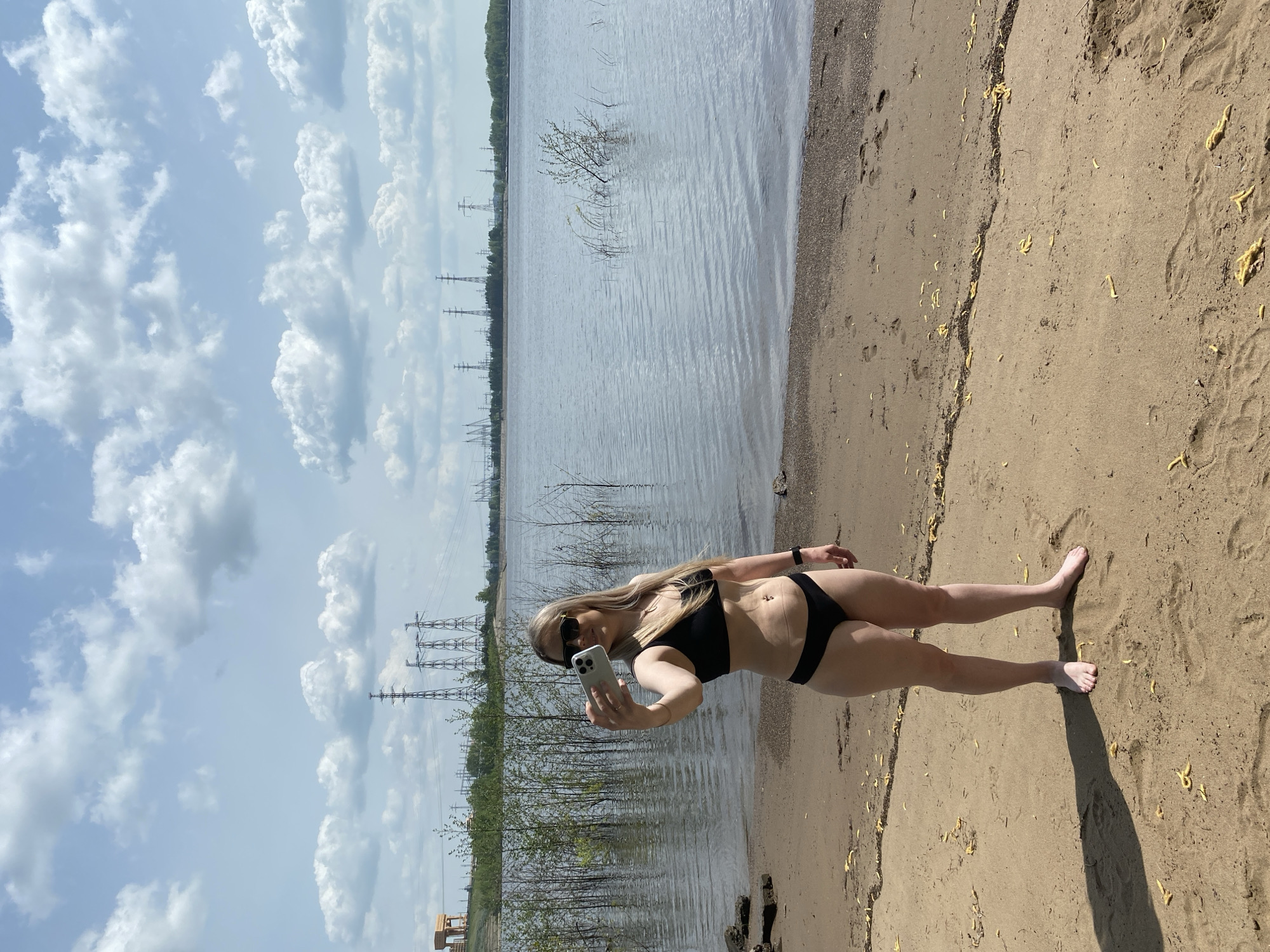 A blonde woman in a black bikini and sunglasses stands on a sandy beach, holding a smartphone and taking a selfie. Behind her is a body of water with trees and vegetation along the shore. In the distance, power line towers are visible. The sky is blue with puffy white clouds. The location is Chaikovsky, Russia.