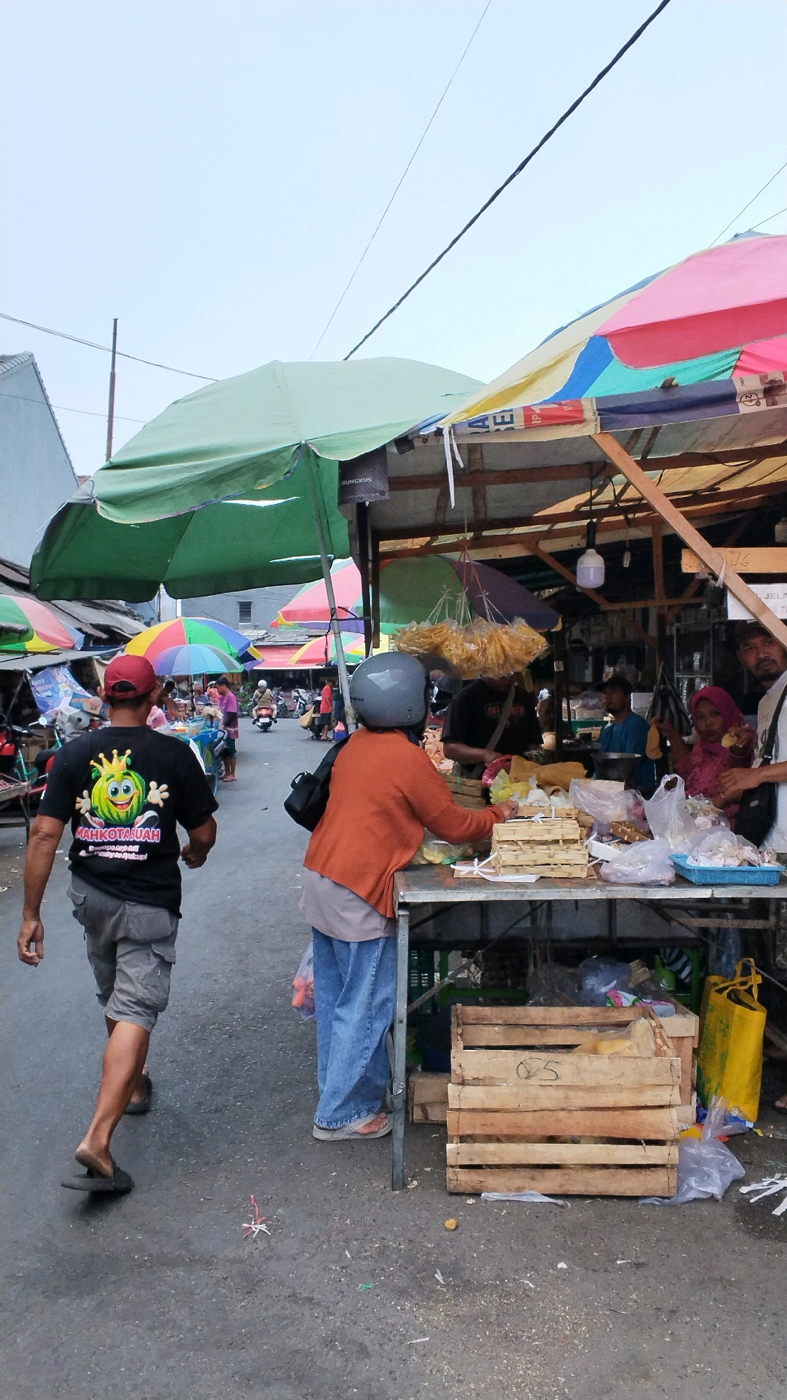 In the city of Jagalan, Indonesia, an outdoor market scene unfolds under a cloudy sky. A man in a black t-shirt with a cartoon watermelon graphic walks away from the camera, his grey cargo shorts and sandals visible. To his right, a woman wearing a bright orange cardigan and blue jeans stands at a market stall, her grey helmet on her head. She is interacting with the stall owner, a person partially obscured behind a table laden with various goods, including what appears to be food items in plastic bags and wooden crates. Behind the stall, several other individuals are visible, one wearing a pink headscarf. Various colorful umbrellas are set up to provide shade, including a large green one that dominates the left side of the frame and a series of smaller, multi-colored umbrellas further in the background. The market appears to be situated on an asphalt road, with more stalls and activity visible in the distance, including people on motorcycles. The ground is littered with some debris.