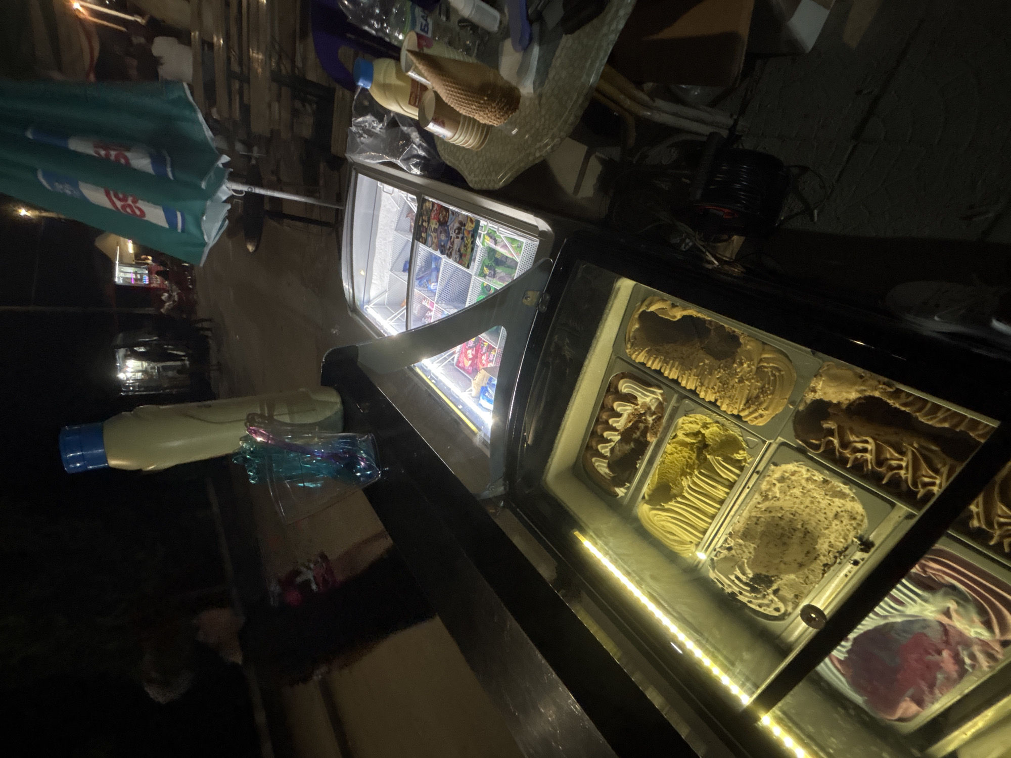 The image depicts an outdoor vendor stand at night, described as being in Kostievo, Bulgaria. In the immediate foreground, a black counter surface is visible, upon which stands a tall, light-yellow plastic bottle with a blue cap and a clear container holding multiple colorful plastic scoops. Directly below the counter is a glass-fronted display freezer containing six large metal trays of various ice cream or gelato flavors, including chocolate, vanilla, a speckled light brown, a ridged yellow, and a pink/red swirl. An LED strip illuminates the interior of this freezer. To the right and slightly behind, a second, smaller glass-fronted freezer displays numerous pre-packaged frozen items, likely ice cream bars or popsicles, arranged in baskets.
To the right of the freezers, a round, translucent table with a textured base holds a stack of brown paper cups, a large textured conical object, and a clear plastic bottle with a partially visible Cyrillic label ("БА"). A dark green market umbrella, partially folded, stands behind the table, displaying a white logo with two stylized 'e's. A dark purple plastic chair is partially visible near the table. On the ground beneath the table, a black electrical cable reel is present. In the background to the left, a person wearing a dark shirt with a colorful shoulder design is partially visible and blurred. The overall background features a dimly lit street or market area with other distant stalls or establishments marked by artificial lights, and several indistinct figures of people suggesting a populated environment.