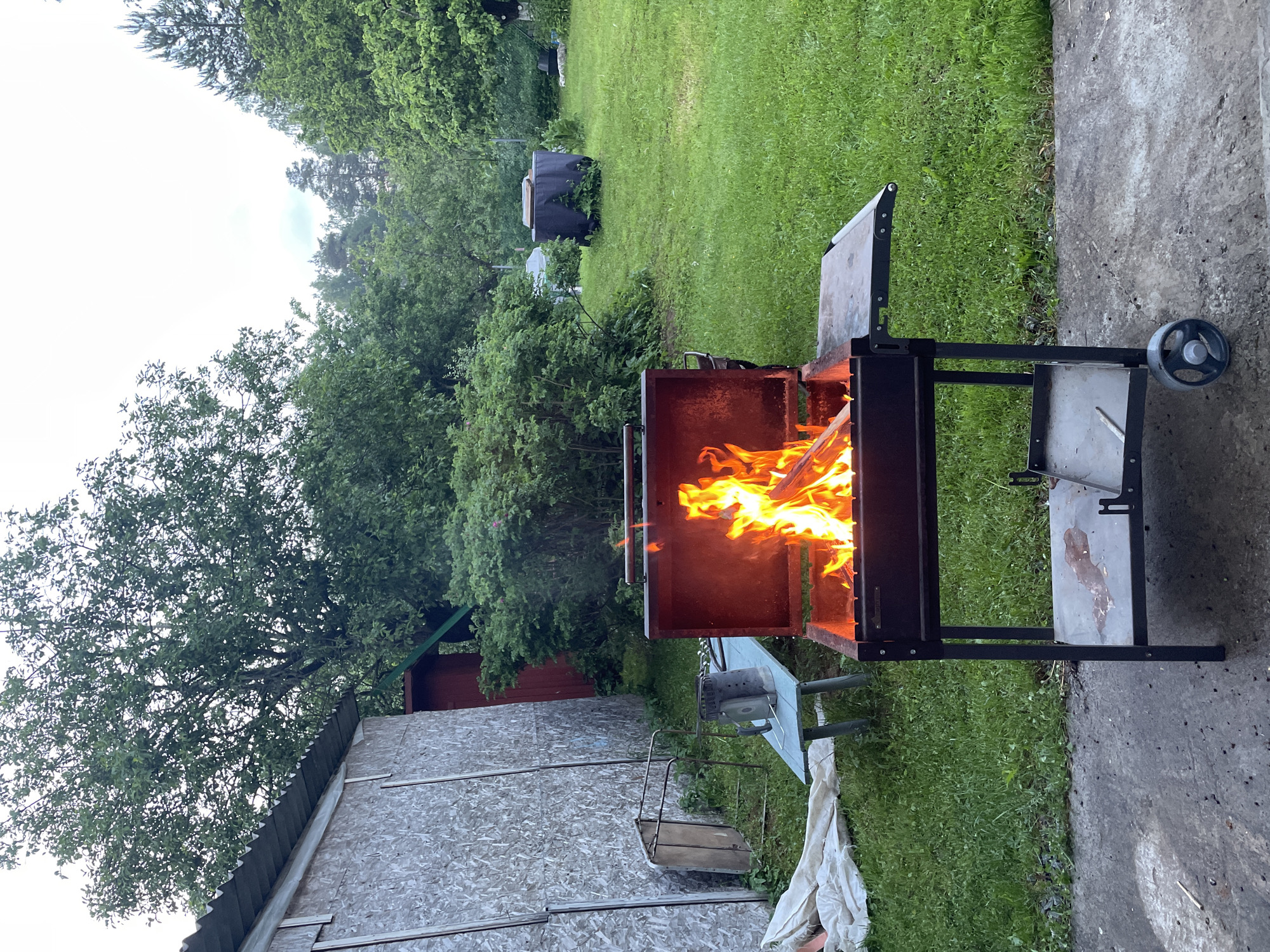 The image depicts an outdoor scene in Sosnovo, Russia, featuring a barbecue grill with active flames. The grill, a portable metal unit with a dark exterior and a red interior, is positioned on a concrete surface. A metal grate with a lower shelf is visible below the main compartment. The grill is situated on a lawn, with lush green grass covering the ground.
In the background, a rustic wooden structure with a corrugated metal roof stands to the left. Adjacent to it is a red shed. Further into the background, dense foliage and trees create a natural backdrop. A light blue metal table with a metal chair is set up near the wooden structure, and a charcoal chimney starter sits on the table. A black cylindrical object, possibly a water tank or barrel, is visible in the distance on the grass. The sky above is overcast. The overall impression is that of a casual outdoor gathering or a preparation for a barbecue.