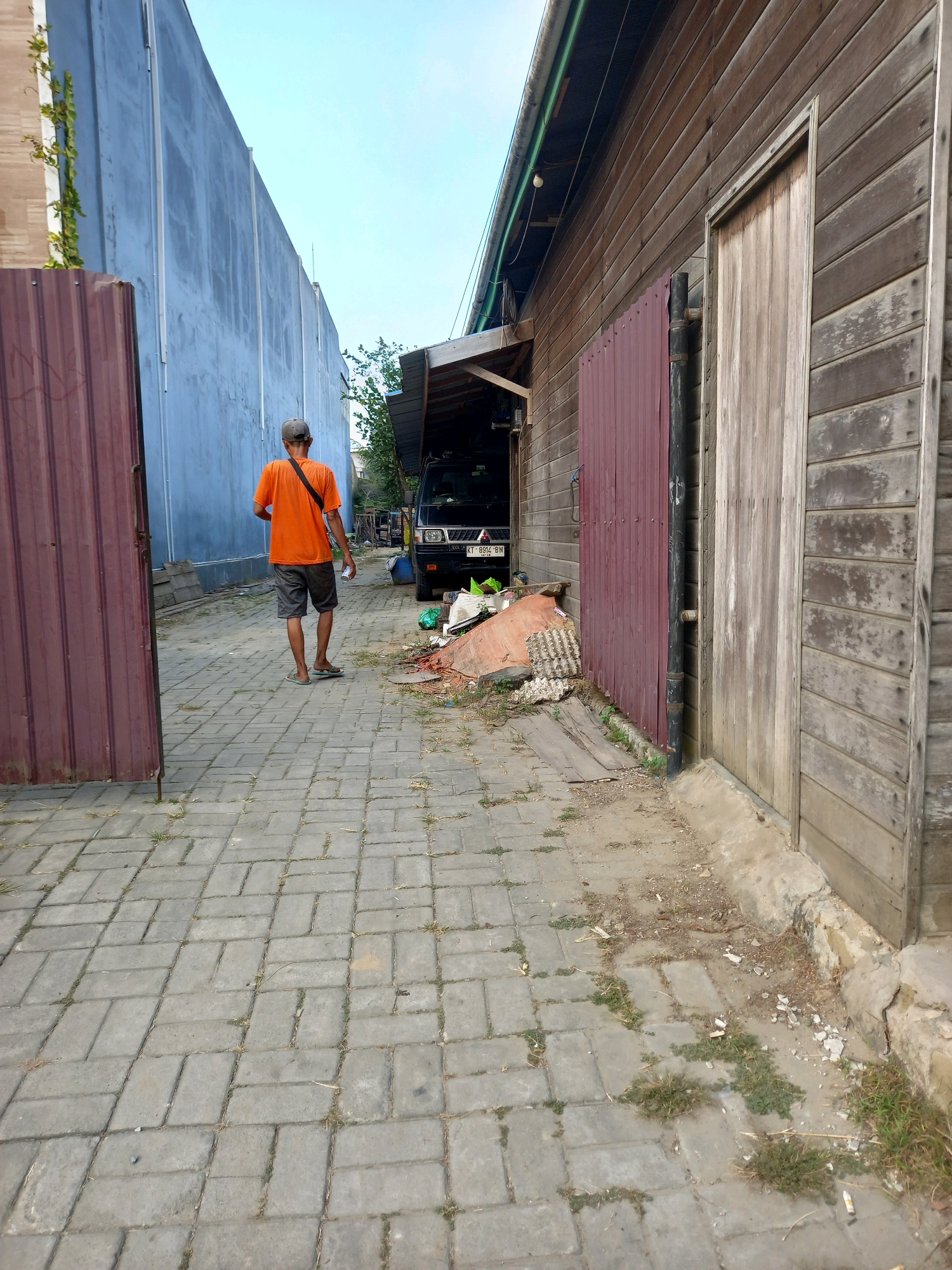 The image shows a man wearing an orange t-shirt and grey shorts walking on a paved walkway. He is carrying a white cup in his right hand and has a black strap across his chest. The walkway is bordered by a tall blue wall on the left and a wooden building with a weathered door on the right. There is debris on the ground near the wooden building, including discarded materials and some vegetation. A black van is parked in the background. The scene appears to be in a semi-urban or industrial area. The license plate on the van, "KT 8914 BW", indicates it is registered in Kalimantan Timur, which includes the city of Samarinda, Indonesia.