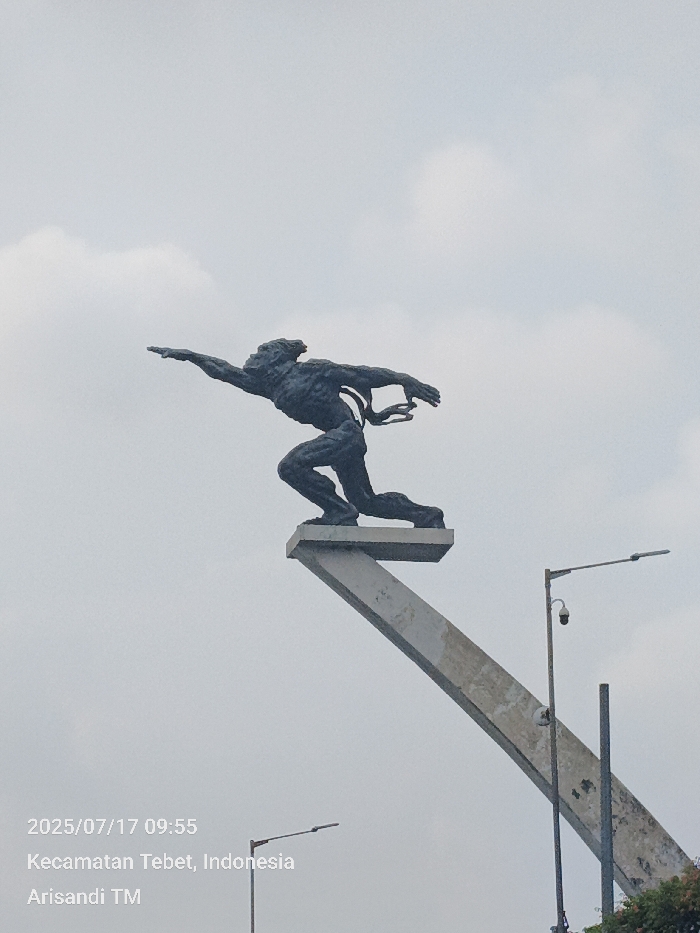 The image depicts a large, dark-patinated bronze statue of a male figure in a dynamic, forward-striding pose, mounted on a light-colored, upward-sloping concrete pedestal. The figure's left arm is extended straight forward, and its right arm is bent with the hand near the chest, with flowing elements trailing behind its back. The background consists of an overcast sky with scattered light gray clouds. To the right of the statue's base stands a tall metal street light pole with a downward-angled lamp and a spherical CCTV camera mounted below it. A second street light pole is partially visible in the lower left, and green foliage is present in the bottom right corner. The image includes a timestamp of 2025/07/17 09:55 and is located in Kecamatan Tebet, Special Capital Region of Jakarta, Indonesia. No individuals are visible.