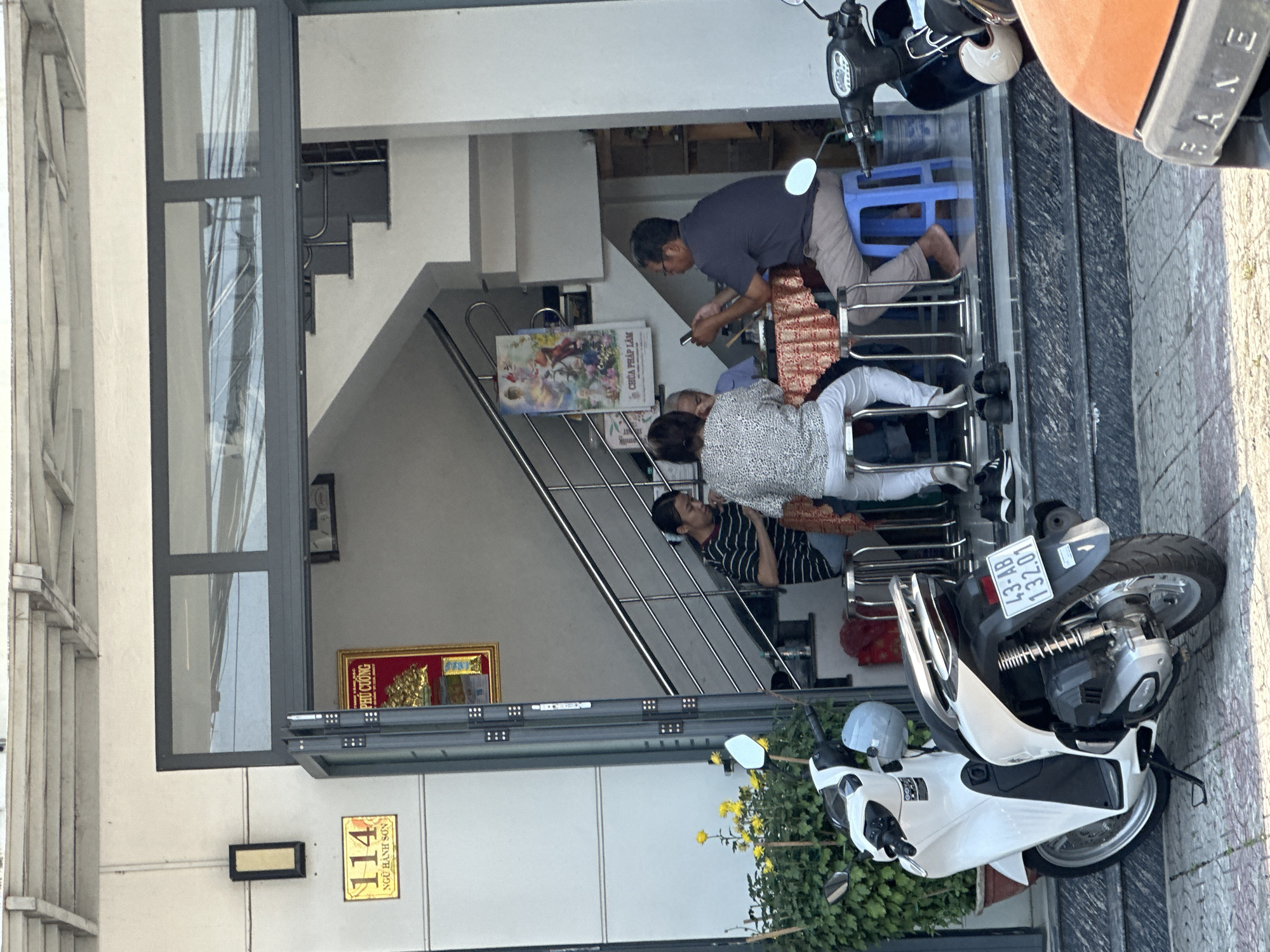 The image shows the facade of a building in Đà Nẵng, Vietnam, with a family gathering visible inside. The building has a large glass-paneled entrance, with stairs leading up inside. A sign on the building reads "114 NGŨ HÀNH SƠN," indicating the address, along with "PHÚ CƯỜNG" above a framed picture. A man is seen crouching, possibly using a mobile phone, while two women and another man are sitting in the foreground. Two motorcycles are parked in front of the building, partially obscuring the view, with one having a license plate reading "43-AB 132.01." A poster with the text "CHÚA PHÁP LÂM" is placed on the stairs. Yellow flowers and greenery are present near the base of the building.