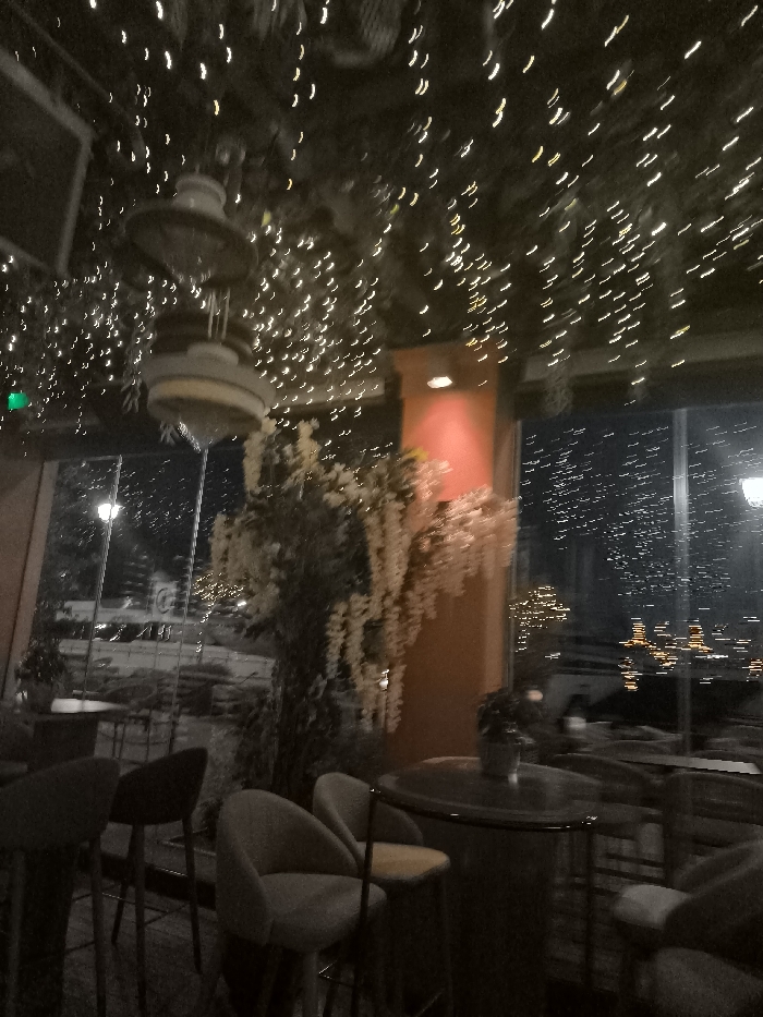 This image captures the inviting interior of an establishment, likely a restaurant, bar, or cafe, at night. The ceiling is a canopy of numerous small, warm-toned string lights, creating a sparkling, magical effect, intermingled with dark foliage. Two large, ornate hanging fixtures, possibly decorative lights or sculptures, are visible in the upper center.
In the mid-ground, a prominent, tall decorative plant with abundant light-colored, drooping flowers stands alongside a reddish-orange pillar that is softly illuminated by an upward-facing light. The foreground features several modern, upholstered chairs or stools with light-colored seats and dark frames, arranged around a round, dark-toned table on which a small potted plant rests.
A large glass window dominates the background, revealing the outside world at night. Through the window, streaks of distant artificial lights, including streetlights and what appear to be urban or possibly coastal lights, are visible, creating a blurred, dynamic backdrop. No individuals are present in the scene. The overall atmosphere is cozy and warm, achieved through the abundant use of decorative lighting against the dark setting. The image has a soft blur, suggesting low light conditions or camera motion. There is no clearly readable text visible. The view through the window hints at an urban or coastal location, consistent with Palaio Faliro, Greece.