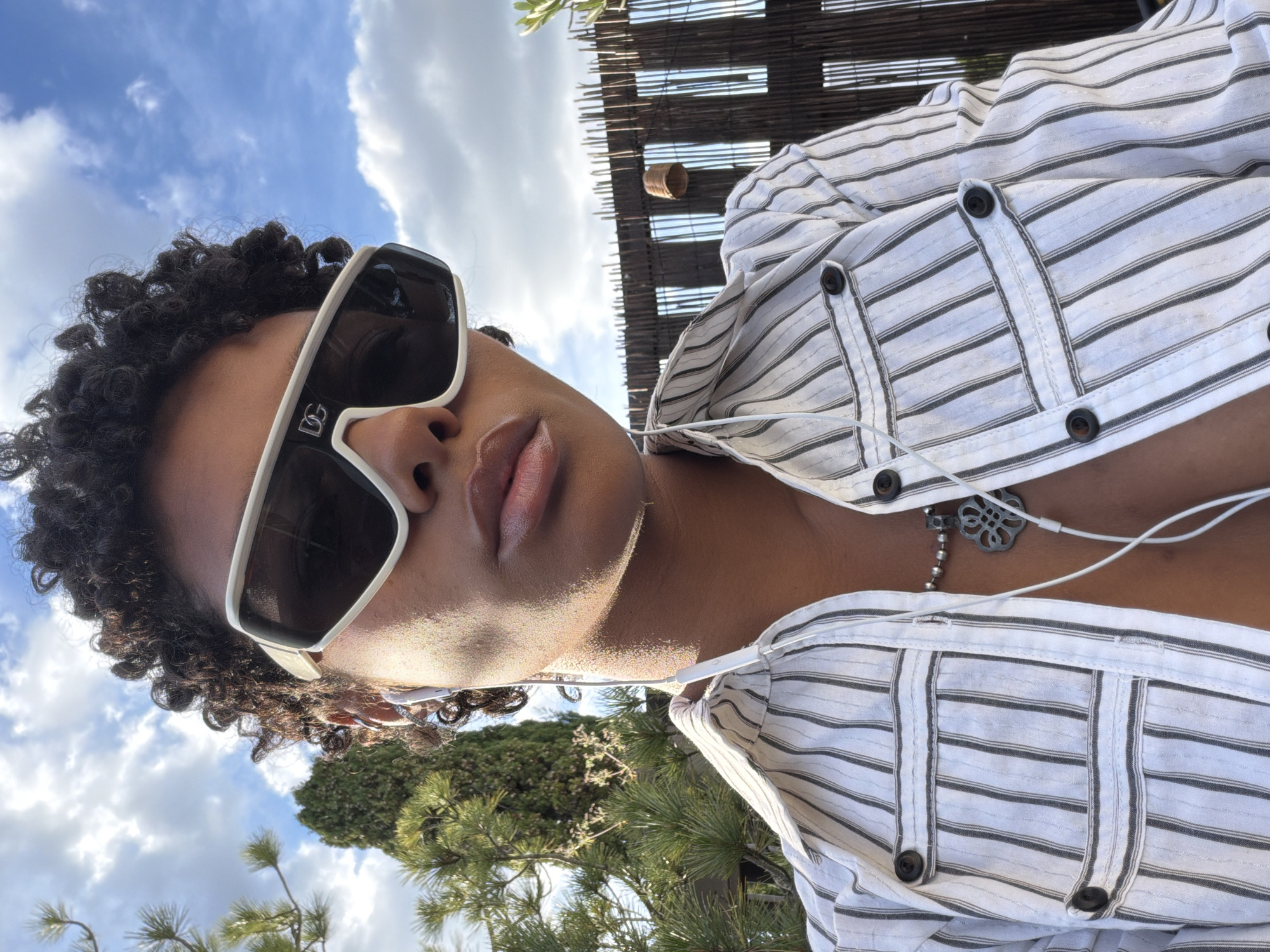 This rotated, upward-facing shot captures a person relaxing outdoors on a sunny day in Champs-sur-Marne, France. The subject, who has dark curly hair, is wearing large white-framed sunglasses with dark lenses. The "DG" logo is clearly visible on the left temple of the sunglasses. Their lips are slightly parted, showcasing a soft, natural lip color.
They are dressed in a white shirt with thin dark horizontal stripes, featuring dark buttons and distinct vertical stitched panels. White wired earphones hang around their neck, draping over the shirt, and a silver necklace with an intricate, possibly quatrefoil-shaped, pendant is visible.
The background features a bright blue sky dotted with scattered white clouds, suggesting clear weather. Green foliage, including visible pine-like trees, and a wooden slat structure, possibly a pergola or sunshade, are also part of the outdoor setting. The bright sunlight casts highlights on the person's skin and hair, emphasizing the daytime scene. The overall impression is one of casual relaxation in an inviting outdoor environment.