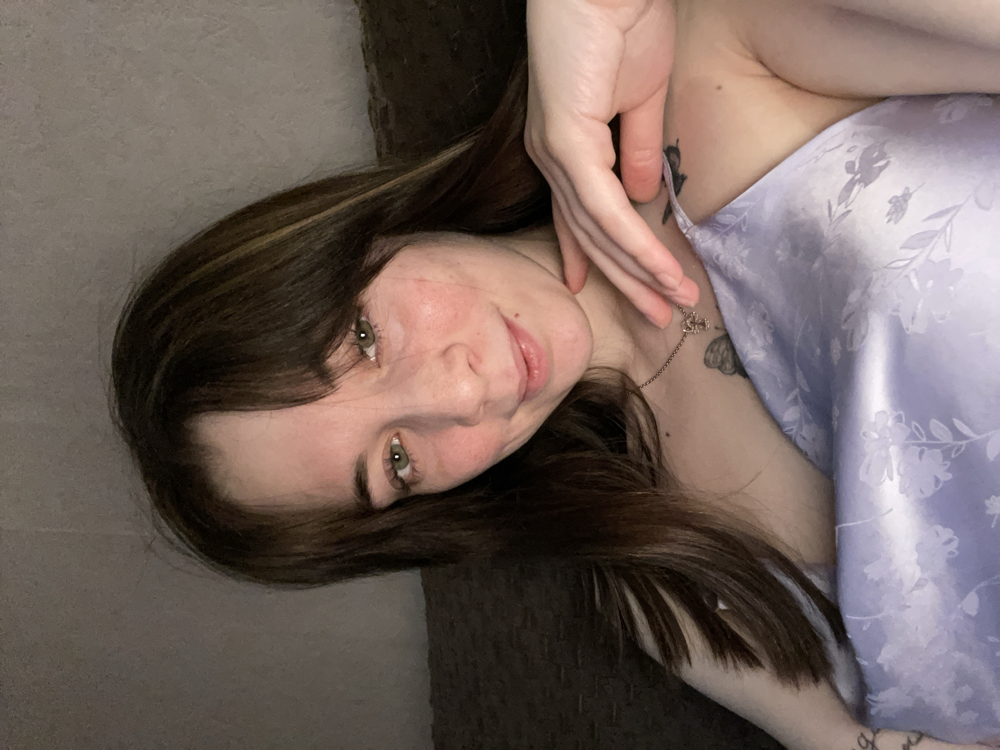 The image shows a young woman indoors. She has dark brown hair with blonde highlights and green eyes. Her skin has some redness. She is wearing a light purple floral-patterned top. A delicate necklace is visible on her neck. She has tattoos on her upper chest and arm. Her hand is near her face. The background shows a dark textured surface. There is no evidence to indicate the location is Chaikovsky, Russia.