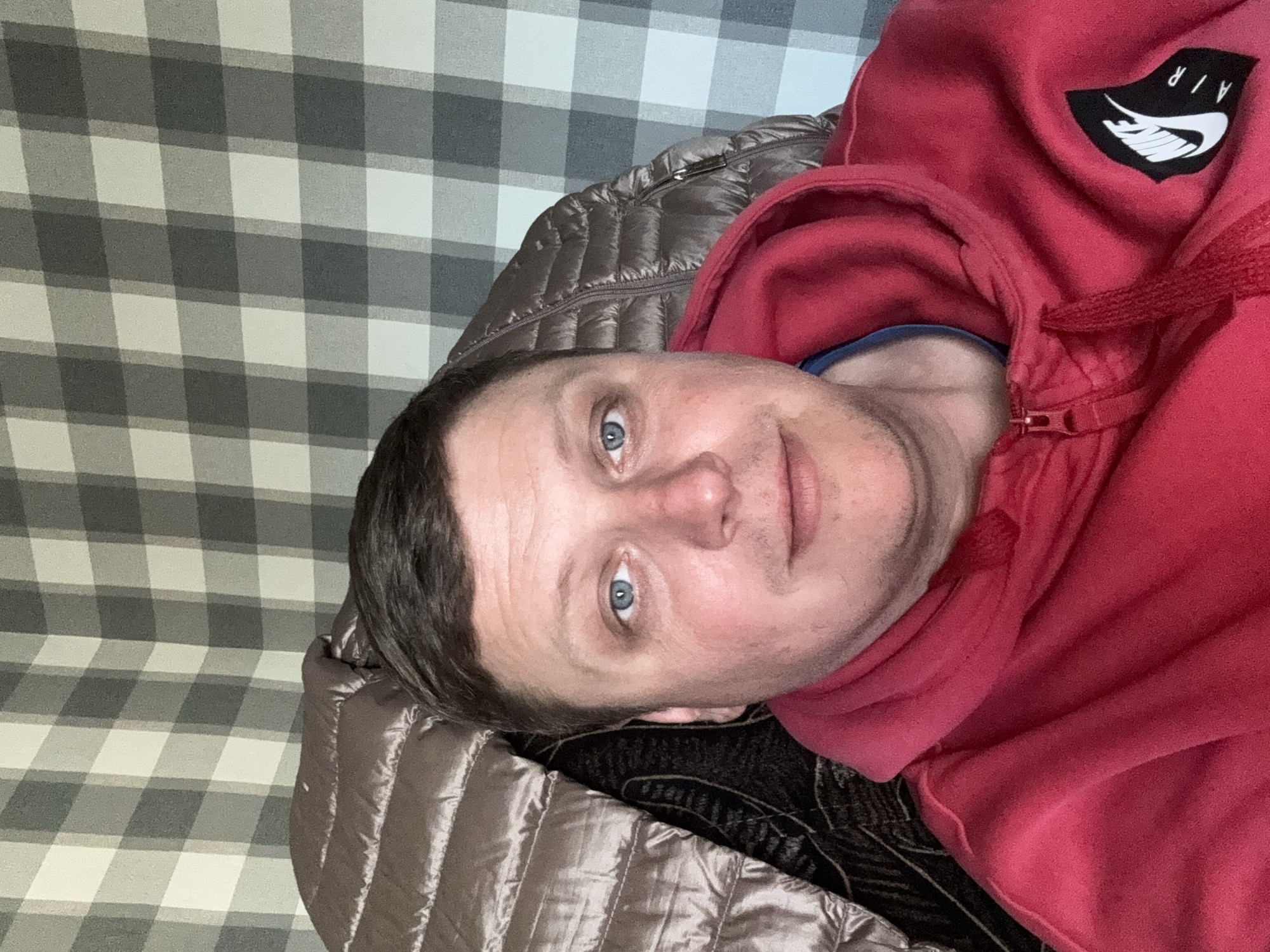 The image shows a person lying down, possibly on a bed or couch, wearing a red hooded sweatshirt with a logo on the upper right side. The person has short, dark hair and blue eyes, looking directly at the camera. The background includes a gray and white checkered pattern, possibly a wall or headboard, and a brown quilted fabric. The location is in the city of Unknown, Unknown.