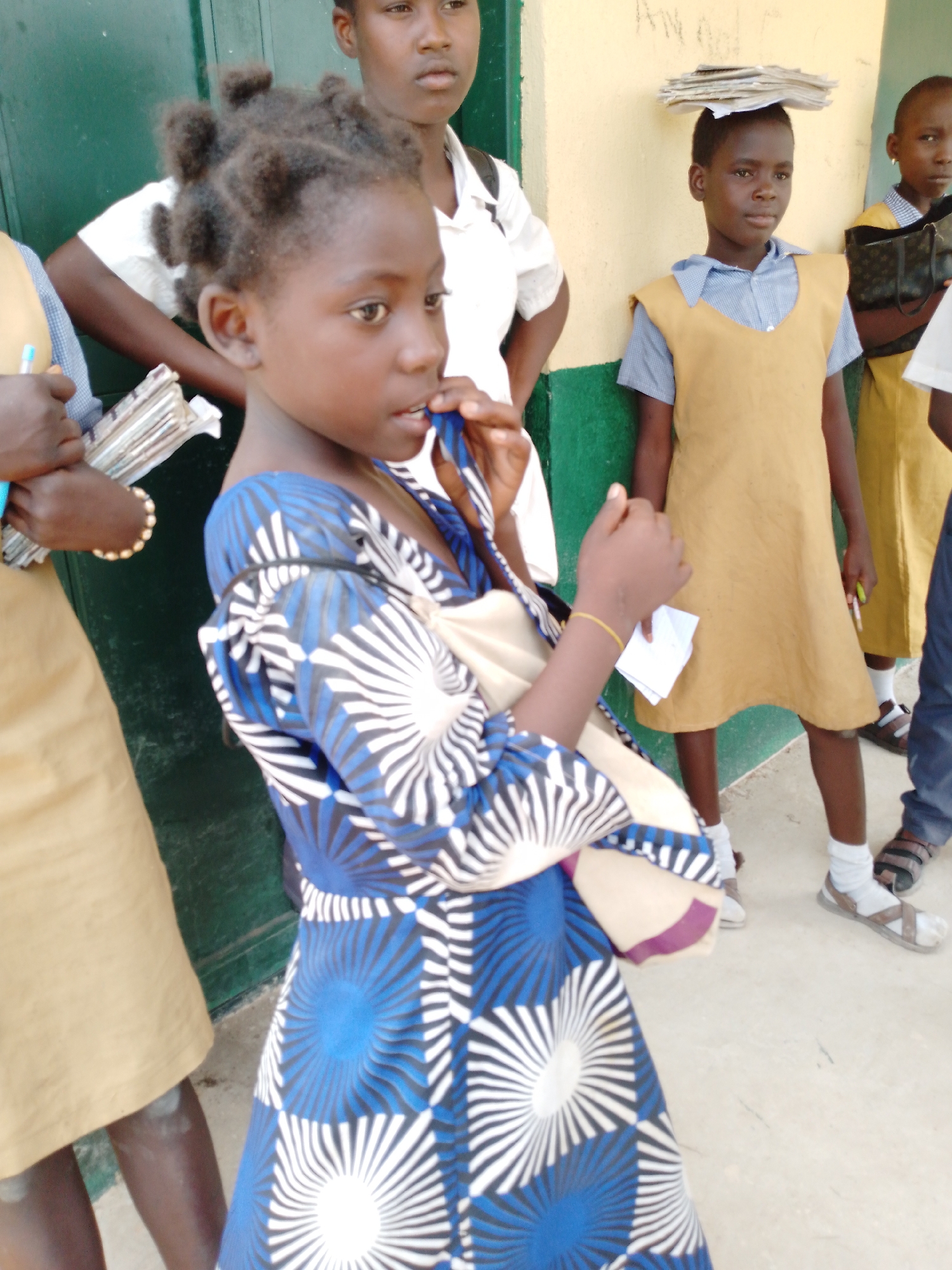 Akwanga, Nigeria: Several African children stand on a light-colored surface, possibly a sidewalk or concrete area. One child is in the foreground wearing a blue and white patterned dress. To her left, another child wears a tan dress and holds papers and a pen. Behind them is a green wall. Another child stands with a stack of papers balanced on her head and wears a tan dress over a collared shirt. There is a tan wall behind her. A girl in a white shirt and tan dress stands behind the girl in the blue and white dress.