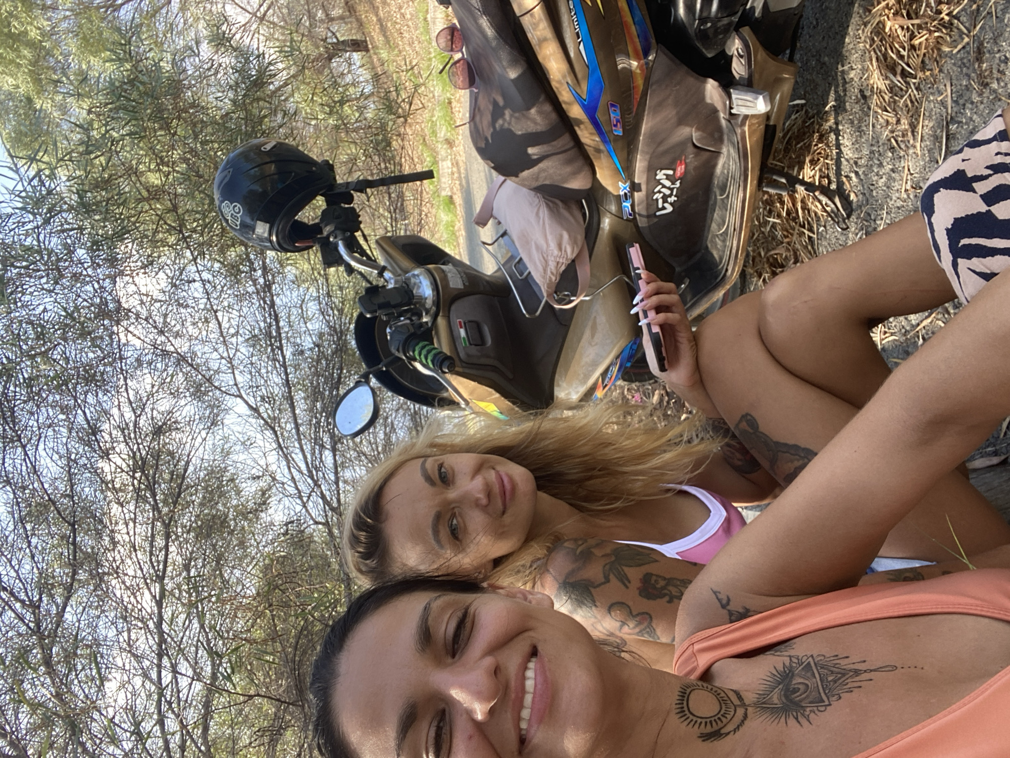 Two women are seated next to a scooter outdoors. The woman on the left, in the foreground, is wearing an orange top and has tattoos on her chest and neck. She is smiling at the camera. The woman on the right is wearing a pink bikini top and has blonde hair. She is looking towards the camera and smiling. The scooter is tan with blue and yellow accents and has a black helmet on its handlebars. The background is filled with trees and sunlight. The scene appears to be in a rural or natural setting, possibly a roadside in Phu Thuy Ward, Vietnam.