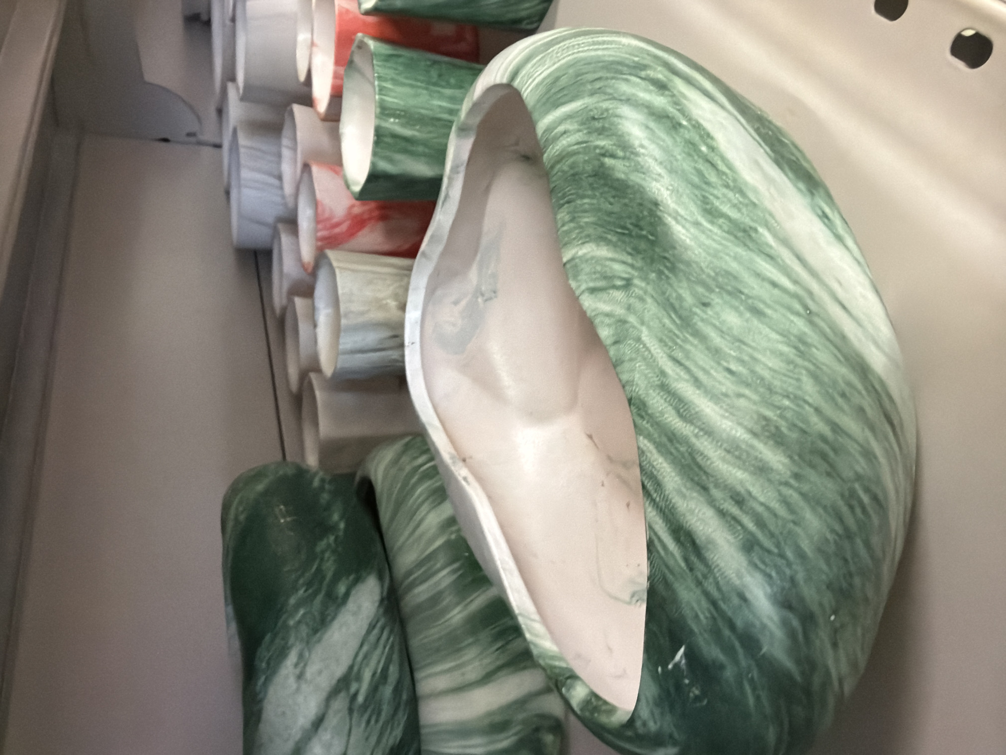 The image shows a collection of decorative ceramic pots with a marbled green and white design. The pots are of various sizes and shapes, with the closest pot being a large, irregular-shaped bowl. In the background, rows of smaller, cylindrical pots with similar marbled patterns in different colors, including pink and white, are visible on shelves. The scene appears to be in a retail setting, possibly a store specializing in home decor or ceramics. The lighting is soft, and the background is out of focus, drawing attention to the pots in the foreground. The image does not contain any information about the city of Pazardzhik, Bulgaria.