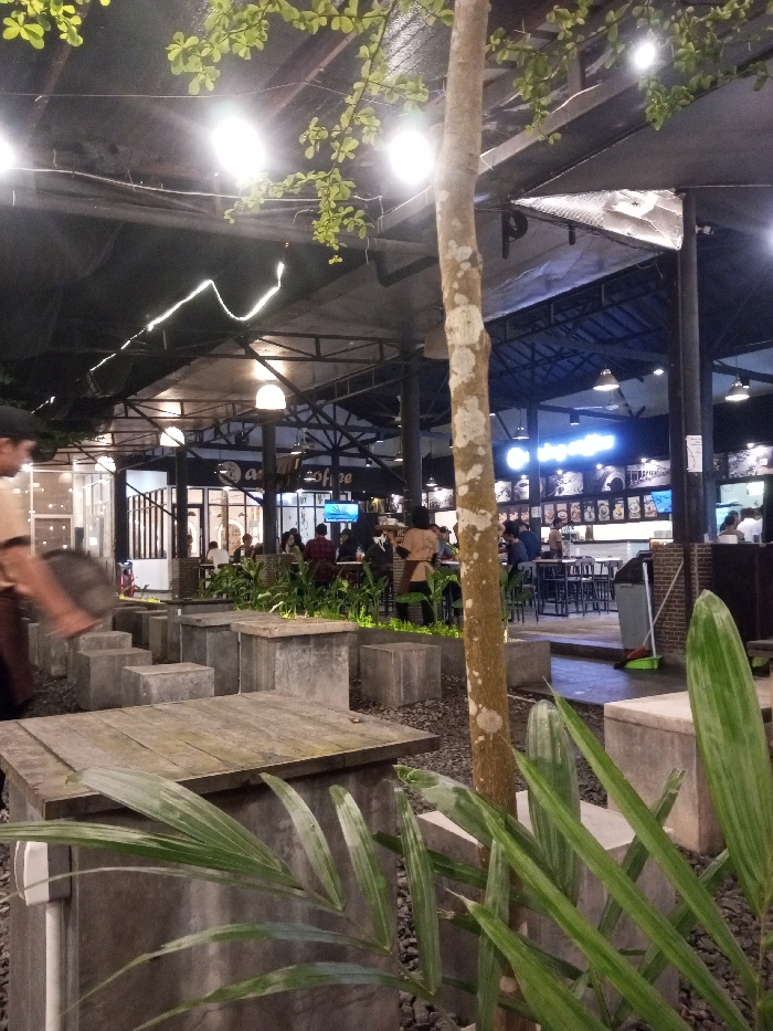 The image displays an illuminated outdoor or semi-outdoor dining area of AR Coffee in Pontianak, Indonesia, observed at night. The ground surface in the foreground and mid-ground consists of dark gravel. Numerous concrete block structures serve as seating and table bases, with some topped by weathered wooden planks. A slender tree trunk with mottled bark stands centrally in the mid-foreground, partially obscured by broad green foliage in the lower right.
On the left, a person, blurred due to motion, is visible from the chest down, wearing a light-colored shirt and dark trousers, moving to the left. In the mid-ground, beyond the gravel seating, multiple patrons are seated at standard dining tables with dark chairs, engaged in various activities typical of a cafe setting. Several individuals are standing or moving near service areas, one wearing a brown apron or vest. Potted green plants are strategically placed among the seating arrangements.
The background features a building with large glass windows and doors, housing the main cafe section. A prominent sign displaying "AR coffee" in white text with blue glowing elements is visible above a service counter area, which features multiple menu boards and display items. Additional patrons are visible at high-top tables in this section. To the far right, a dark trash receptacle and a green broom with a dustpan are positioned against a brick wall. The overhead structure comprises a dark canopy or roof supported by a metal truss framework, from which numerous bright spherical light fixtures, string lights, and utilitarian lamps are suspended, providing the primary illumination for the scene.