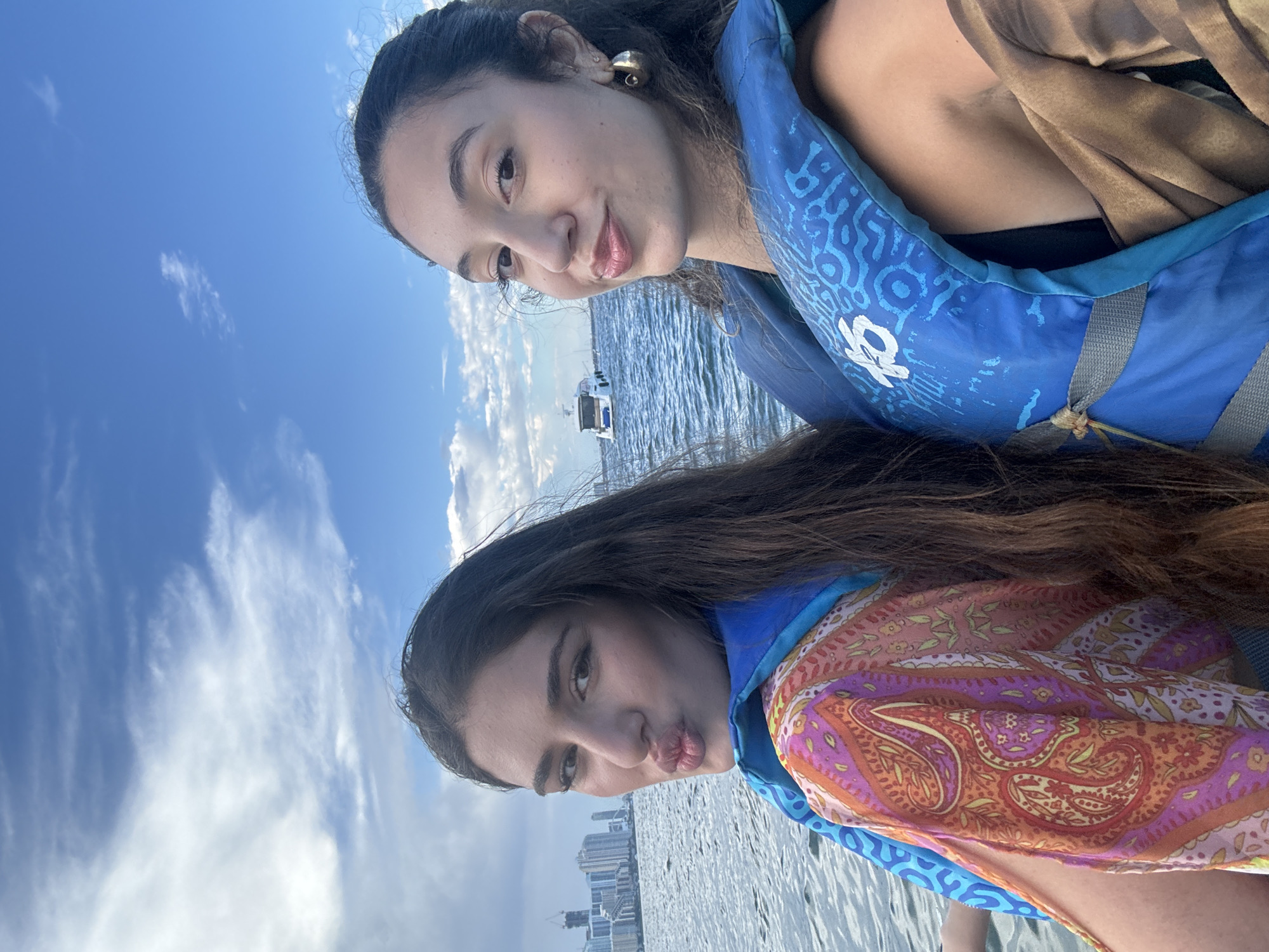 Two young women, wearing blue life vests, are taking a selfie on a boat in the Miami, United States of America. The woman on the left is leaning back and puckering her lips. She is wearing a colorful, patterned top. The woman on the right is looking towards the camera with a slight smile. She is wearing a gold hoop earring. In the background, a city skyline with tall buildings is visible, and a white boat is sailing on the water. The sky is blue with scattered clouds.