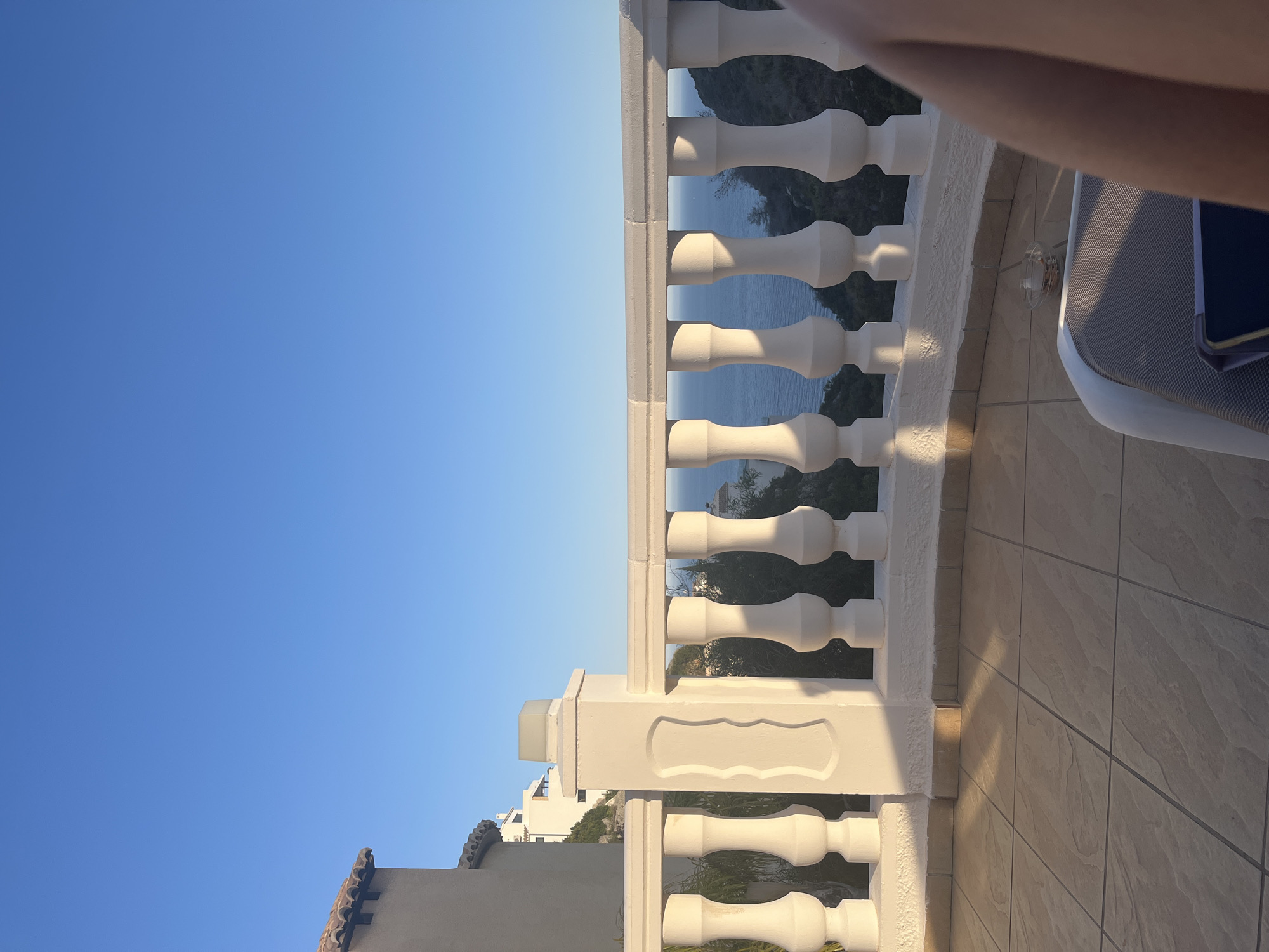 The image shows a view from a balcony in El Poble Nou de Benitatxell/Benitachell, Spain. The balcony features a white balustrade with classic baluster shapes. Behind the balustrade, a glimpse of the ocean and vegetation is visible. A clear blue sky fills the upper portion of the frame. The balcony floor is tiled in light brown with subtle patterns. A section of a white outdoor lounge chair is seen in the upper right corner. A mobile phone is placed on the lounge chair. The image appears to be rotated 90 degrees counter-clockwise. In the lower left, a building with terracotta roof tiles is seen. The balustrade partially obstructs the view of surrounding buildings nestled among the trees.