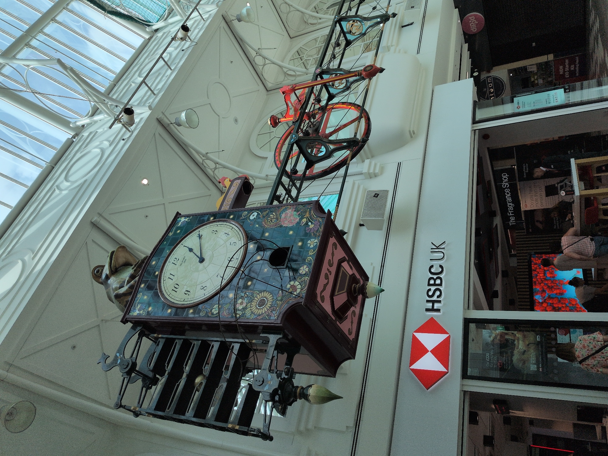 The image shows the interior of a shopping center in Telford, United Kingdom. Dominating the upper portion of the frame is a large, ornate clock with a frog figure at its apex. The clock is suspended from the ceiling and features a dark blue tiled face decorated with floral patterns and Roman numerals. To the right of the clock, a red and black mechanical bicycle is mounted on a railing. Below this, a section of an HSBC UK bank branch is visible, identified by its prominent red and white hexagonal logo and signage. The HSBC entrance has large glass windows displaying promotional materials and interior lighting. Further to the right, partially visible, is a store labeled "The Fragrance Shop." The environment is brightly lit by natural light filtering through a glass ceiling, with white architectural elements and suspended lighting fixtures throughout. Several individuals are present in the lower part of the image, near the entrance to the HSBC bank and the Fragrance Shop, some of whom appear to be customers.