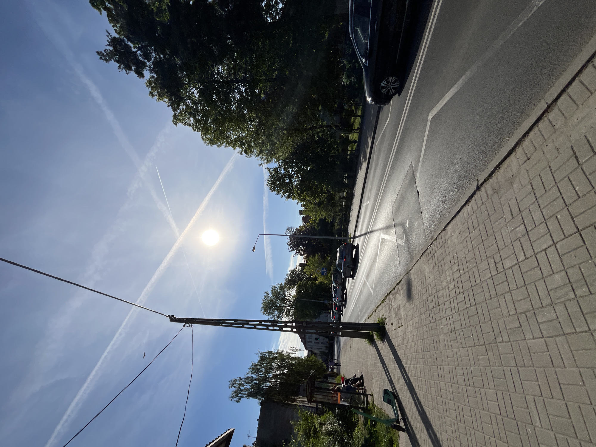 The image depicts a street scene in Unknown, Unknown. A paved sidewalk lines the right side of a road where cars are parked and driving. The sky is visible with a bright sun and contrails. A utility pole is on the left, with wires running across the sky. Trees are visible along the roadside.