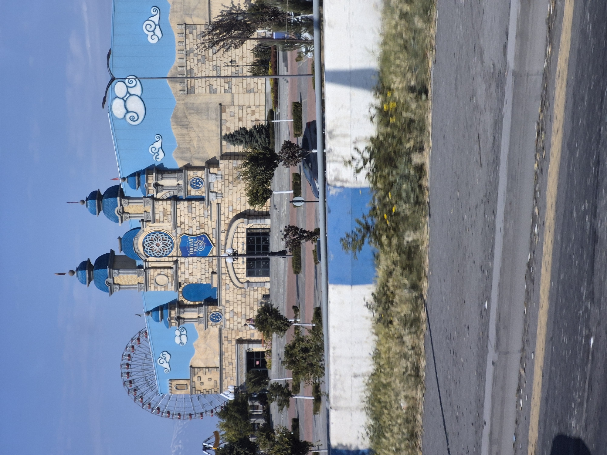 The image depicts Yerevan Park, an amusement park located in Yerevan, Armenia. The park's main building is designed to resemble a castle with blue turrets and accents, set against a backdrop of a blue sky with painted clouds and stylized mountains. A large Ferris wheel is visible behind the main building, to the left. In front of the building, there is a paved plaza with trees, lampposts, and the park's entrance. A grey sedan is parked in the area in front of the building. The foreground shows a road and a decorative barrier with blue and white stripes, separating the road from the park. The overall scene is brightly lit, suggesting a clear day.