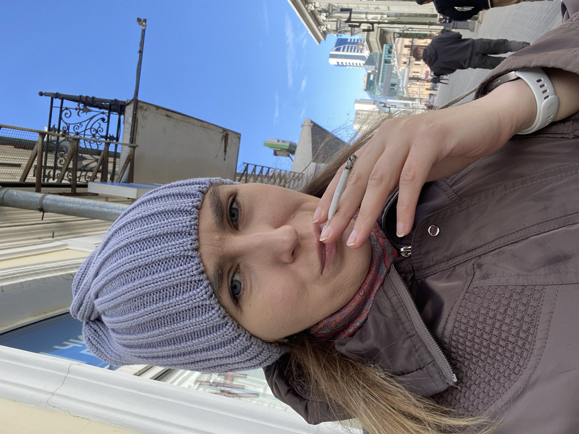 A woman with fair skin, wearing a knitted light purple beanie, a patterned scarf, and a brown jacket, is holding a lit cigarette between her fingers. She is also wearing a white smartwatch. The background shows an urban scene with buildings and a blue sky. There are pedestrians walking in the background. The location appears to be in Yekaterinburg, Russia, based on the urban environment and architectural style of the buildings.
