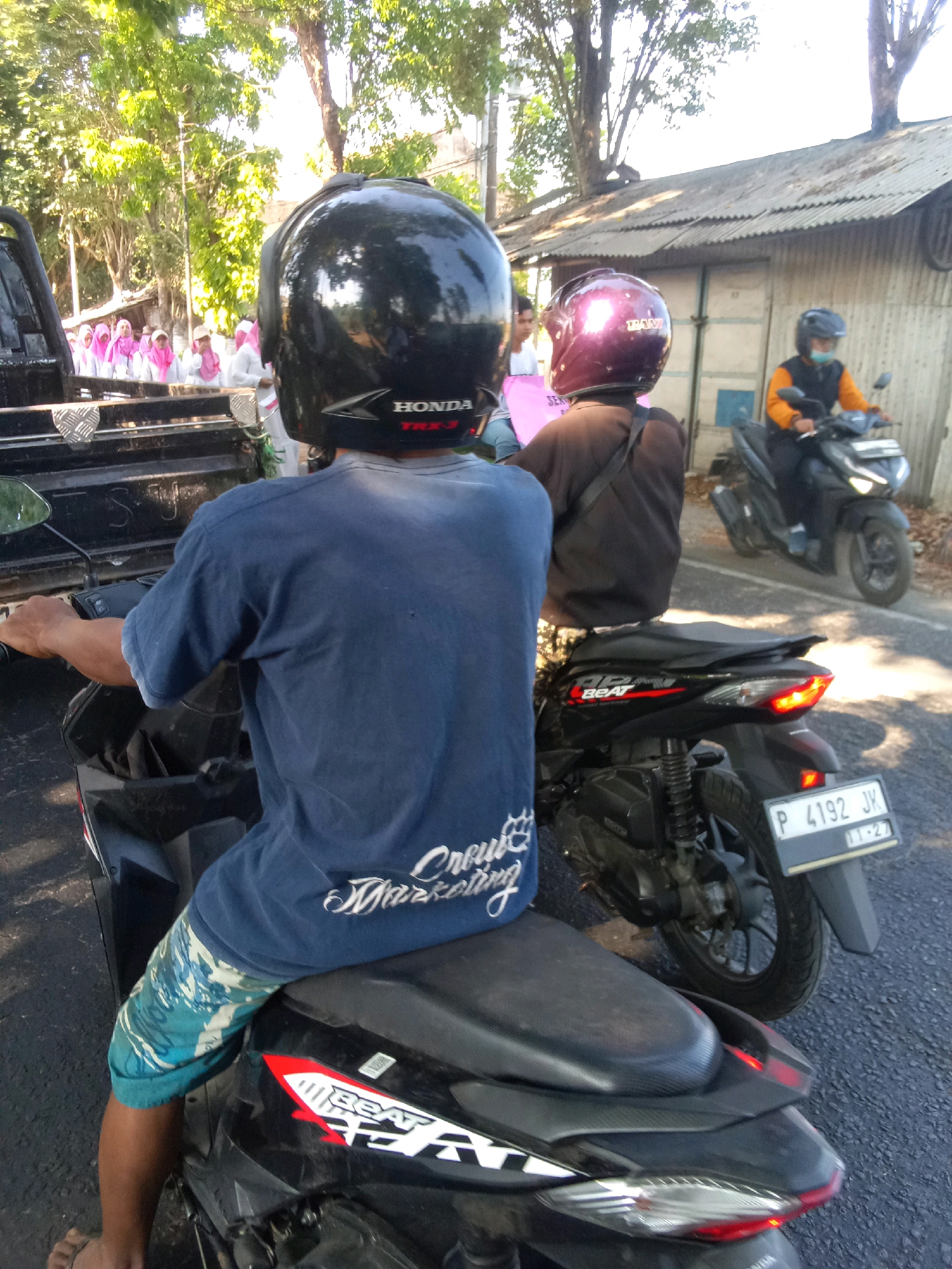 In Pancakarya, Indonesia, two men are riding motorcycles. The man in the foreground, wearing a blue t-shirt and patterned shorts, is riding a black scooter. He has a black helmet with "HONDA TRX-3" written on the back. To his right, a man wearing a black long-sleeved shirt and a maroon helmet is also on a motorcycle. In the background, a group of women wearing pink hijabs and white clothing are standing near a pick-up truck. Another man on a motorcycle, wearing a black vest over an orange shirt and a face mask, is visible further in the background. The scene takes place on a paved road, with trees and a building with a corrugated metal roof visible. The weather appears sunny.