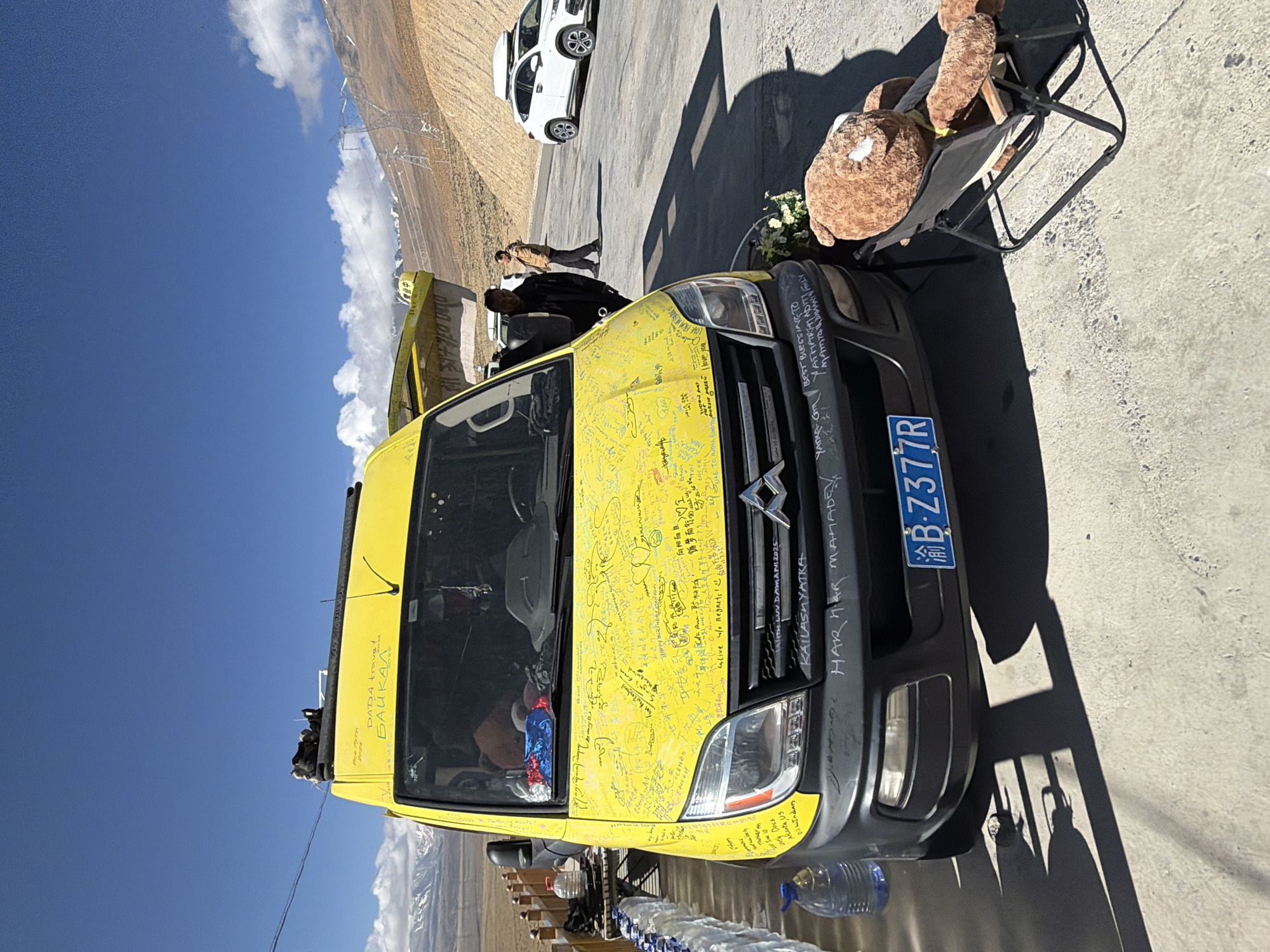 The image displays a vibrant yellow van, likely a Citroën model, parked on an asphalt road in what appears to be a mountainous region of Gyirong County, China. The van is extensively covered in handwritten messages, signatures, and drawings in various languages, suggesting a long journey or pilgrimage. Its license plate, "渝B-Z377R," is clearly visible on the front bumper.
Near the front of the van, a black folding chair holds two brown plush teddy bears, one larger than the other, and several large plastic bottles of water are arranged on the ground. Two individuals are seen standing near the middle of the yellow van, and a white SUV is parked further down the road.
The background reveals a rugged, dry landscape dominated by mountains, with some peaks exhibiting snow caps. Large power lines and transmission towers stretch across the terrain. The sky is clear blue with scattered white clouds, and the bright sunlight casts sharp shadows, indicating midday conditions.
Notable text visible on the van includes "DADA travel BAIKAL," "KAILASH YATRA," "HAR HAR MAHADEV," and phrases like "BEST BLESSING TO" and "live w/o Regrets!". Chinese characters read "平安喜乐国" (Peace and happiness nation) and "向阳而生, 循梦而行" (Born facing the sun, pursuing dreams). Other inscriptions include "Trekker th," "svely Docs Atlanta, Us Dilhi Indian," and names such as "YATHARTAI ADITI FILY MAMTA DOMANI," along with what appears to be "LOVE TO INDIA ROAD TRIP." The overall scene suggests a significant and personal adventure.
