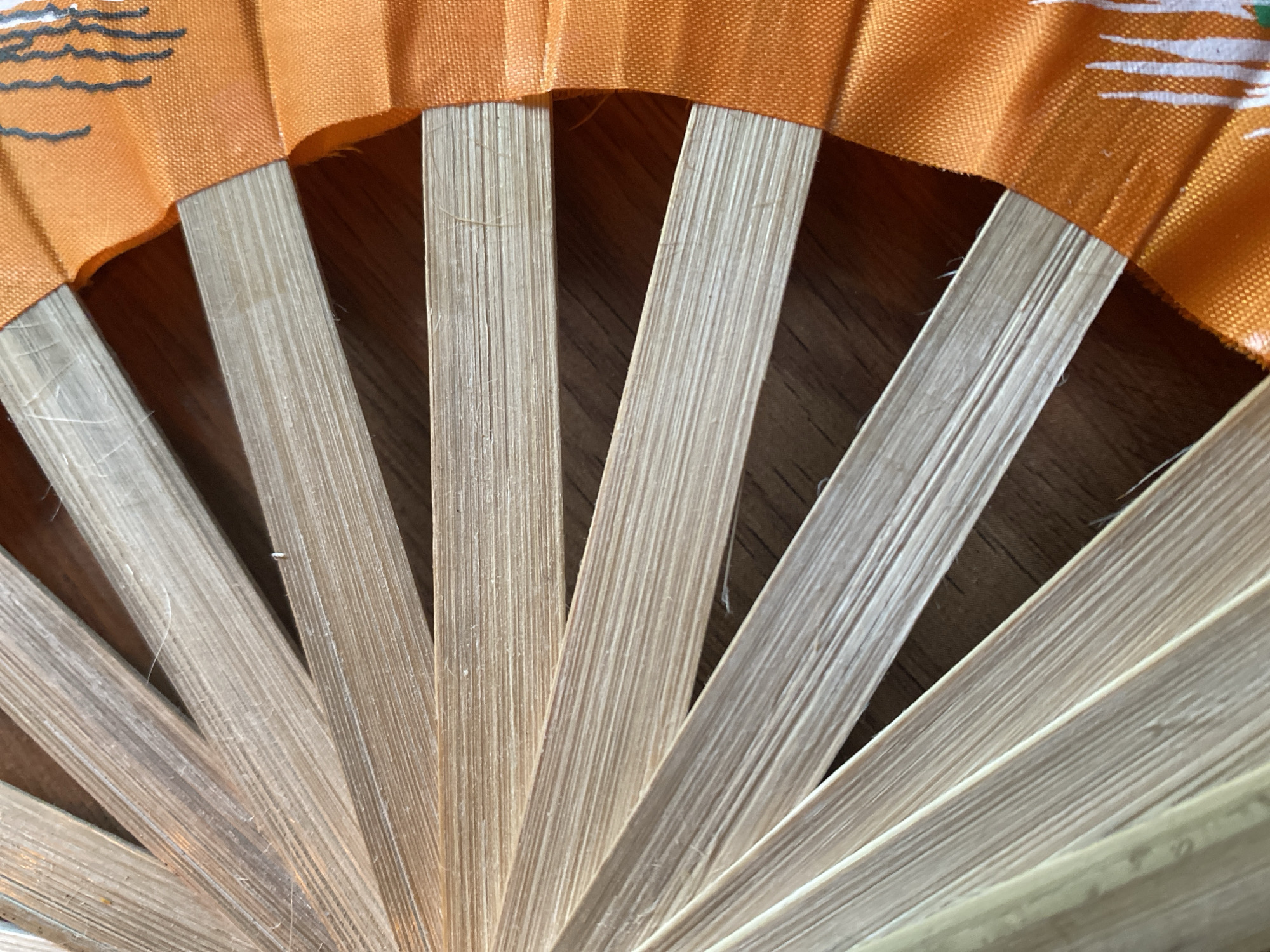 The image shows a close-up of a partially open hand fan. The fan has light-colored, wooden ribs and an orange fabric section at the top edge. The fabric appears to have a simple design with black and white elements. The background is a dark, wood-like surface. The setting doesn't give any indication of location to determine if the setting is Baan Kai, Thailand.