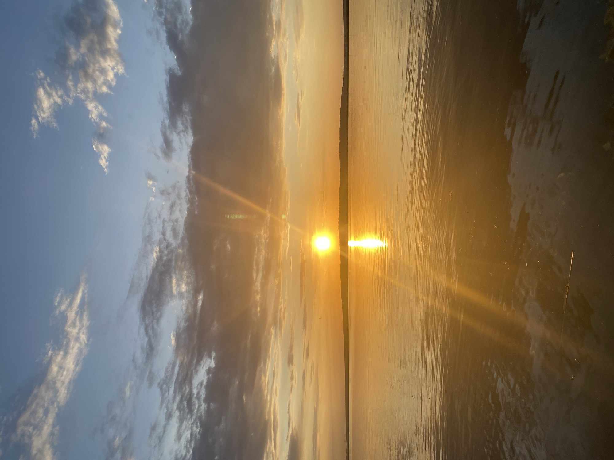 The image shows a sunset over a body of water, possibly a lake or river. The sun is low on the horizon, casting a golden light across the water and the sky. There are clouds in the sky, some of which are illuminated by the setting sun. The far shore is visible as a faint dark line near the center of the image. The exact location is difficult to ascertain without further information.