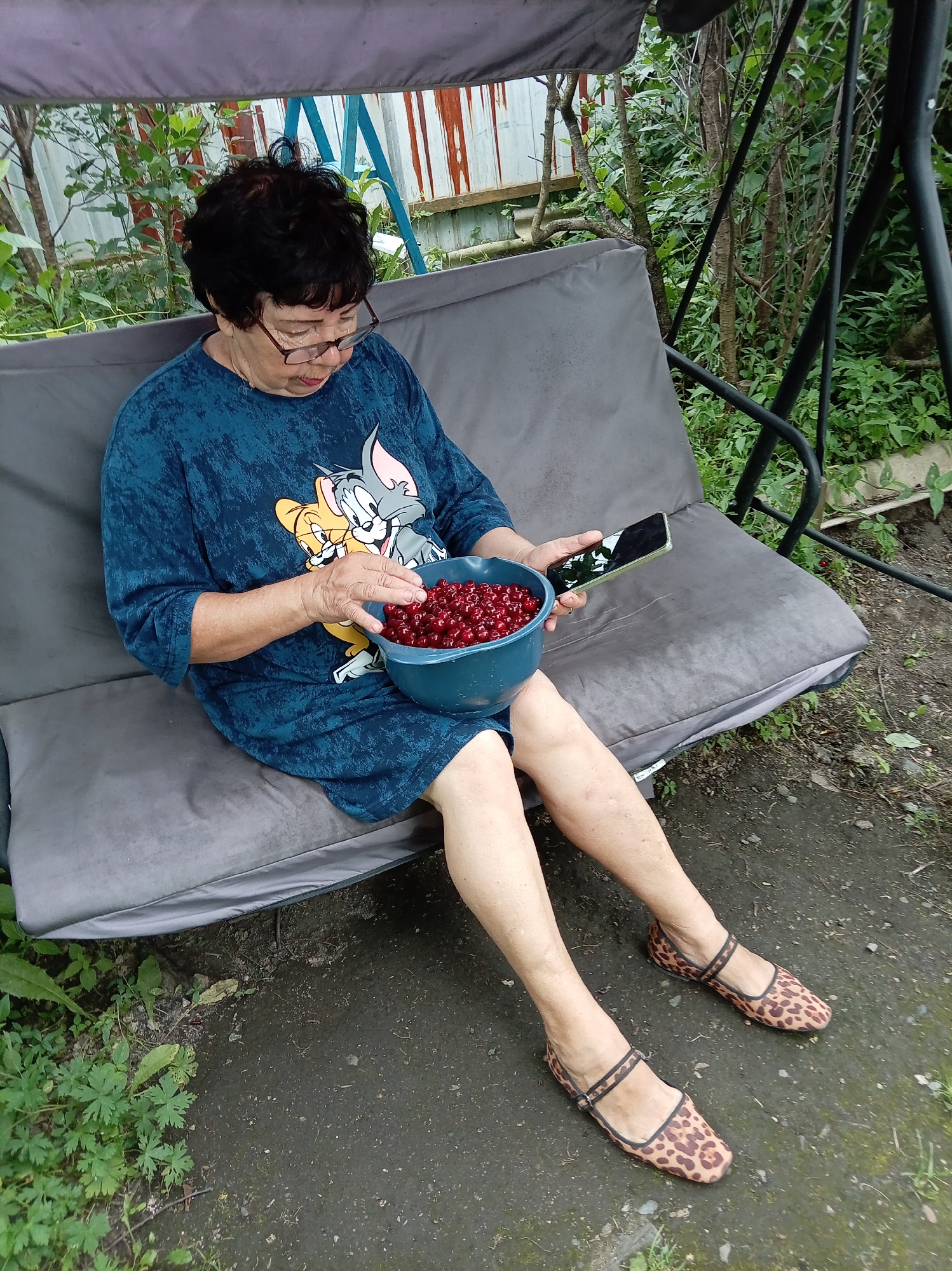 An elderly woman with dark hair and glasses sits on a gray outdoor swing seat, holding a blue bowl filled with dark red cherries in her lap. She is wearing a dark blue t-shirt with a cartoon image of Tom and Jerry on it. In her right hand, she holds a smartphone with a green case. The woman's legs are extended forward, and she is wearing leopard-print ballet flats. The swing is positioned in a yard with green foliage and trees surrounding it, suggesting a natural, possibly rural or suburban setting. The ground appears to be a mixture of dirt and asphalt. The background includes a metal fence and some weathered wooden structures. The scene appears to be outdoors, in daylight, with the overall atmosphere suggesting a moment of relaxation or casual activity. The location is identified as Artyom, Russia.
