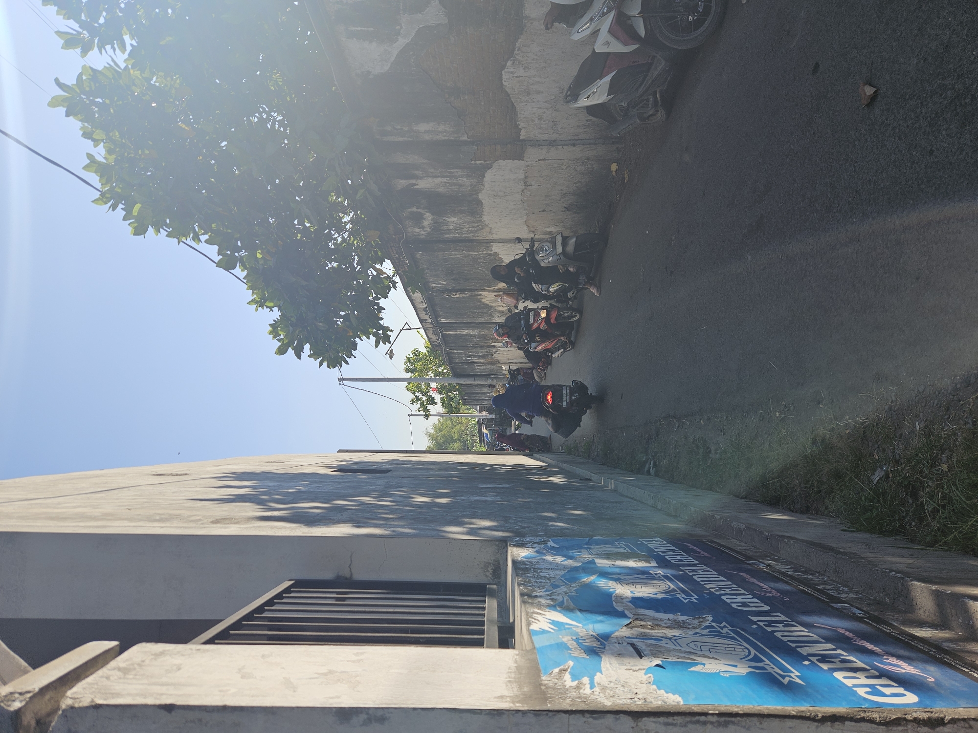 The image depicts a narrow, paved street in a city in Perak, Indonesia. The street is flanked by a concrete wall on the right, with a building wall and a fence on the left. Several motorcycles are parked or being ridden on the street. In the foreground on the left, a portion of a building wall is visible, with peeling blue posters advertising something called "GRENDEL GRENDEL INTERNATIONAL". A dark-colored motorcycle with a rider wearing a dark hijab is in motion, heading away from the camera. Further down the street, other motorcycles and individuals are visible, some appearing to be standing or gathered. A tree with green leaves overhangs the wall on the right, and the sky is clear and blue. The lighting suggests it is daytime, with the sun casting shadows and causing lens flare.