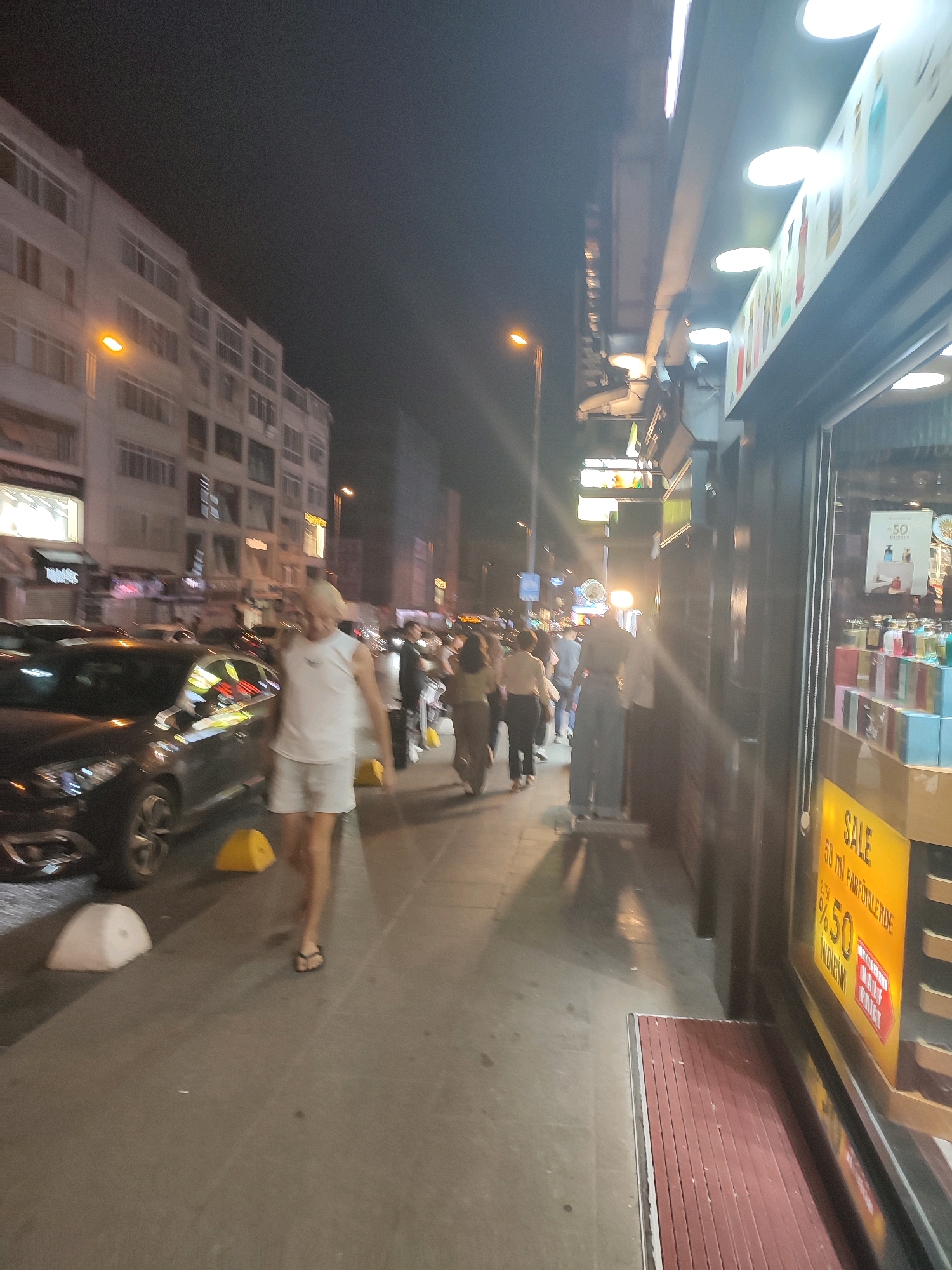The image captures a street scene at night in Kadıköy, Turkey. A person, appearing to be a man with blonde hair, is walking on the sidewalk wearing a white sleeveless shirt and white shorts. He is walking towards the left side of the frame. To his left, several cars are parked along the street. Across the street, a multi-story building with windows is visible. The sidewalk is lined with storefronts. One storefront has a display of perfumes or cosmetics. A large yellow sign with red text indicating "SALE %50" and "50 ml PARFÜMLERDE" is prominently displayed on the storefront. Other pedestrians are walking in the background, some near the storefronts and others further down the street. The lighting suggests it is evening or night.