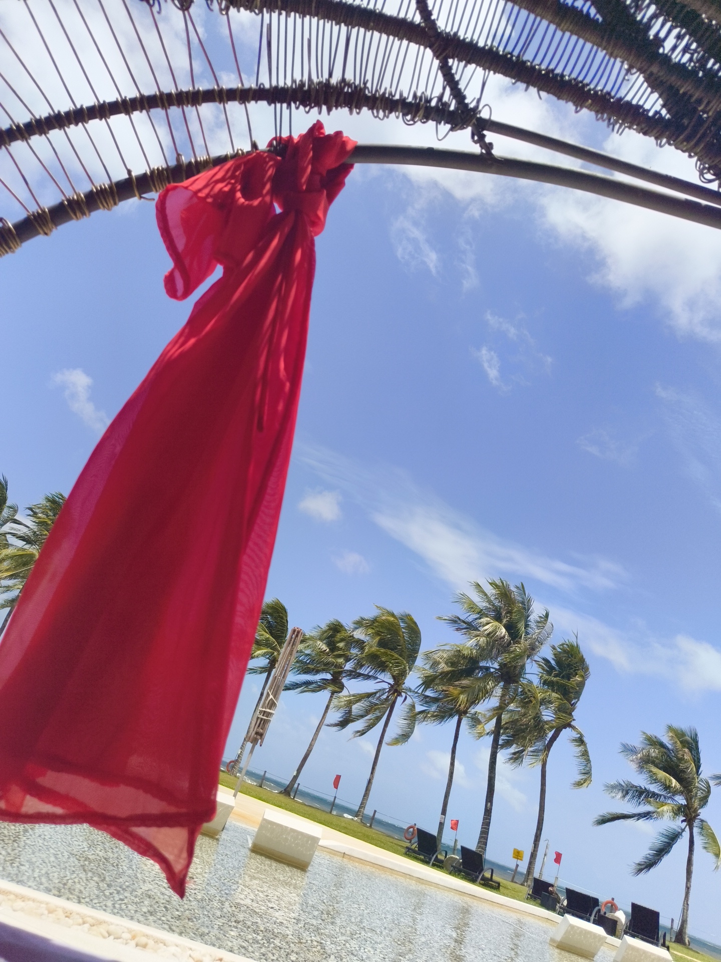 A bright red fabric is tied to a curved, bamboo-like structure, its material billowing in the wind. The fabric is in the foreground, partially obscuring the view of a tropical scene. Beyond it, a clear blue sky with scattered white clouds stretches overhead. A line of palm trees, their fronds rustling in the breeze, are visible in the middle ground, their trunks leaning. In the background, the ocean meets the horizon, with a stretch of green grass and what appears to be lounge chairs near a body of water that could be a pool or part of the beach. Small red flags are planted along the grassy area. The overall impression is one of a resort or coastal vacation spot. The image appears to be taken outdoors, likely during the daytime.