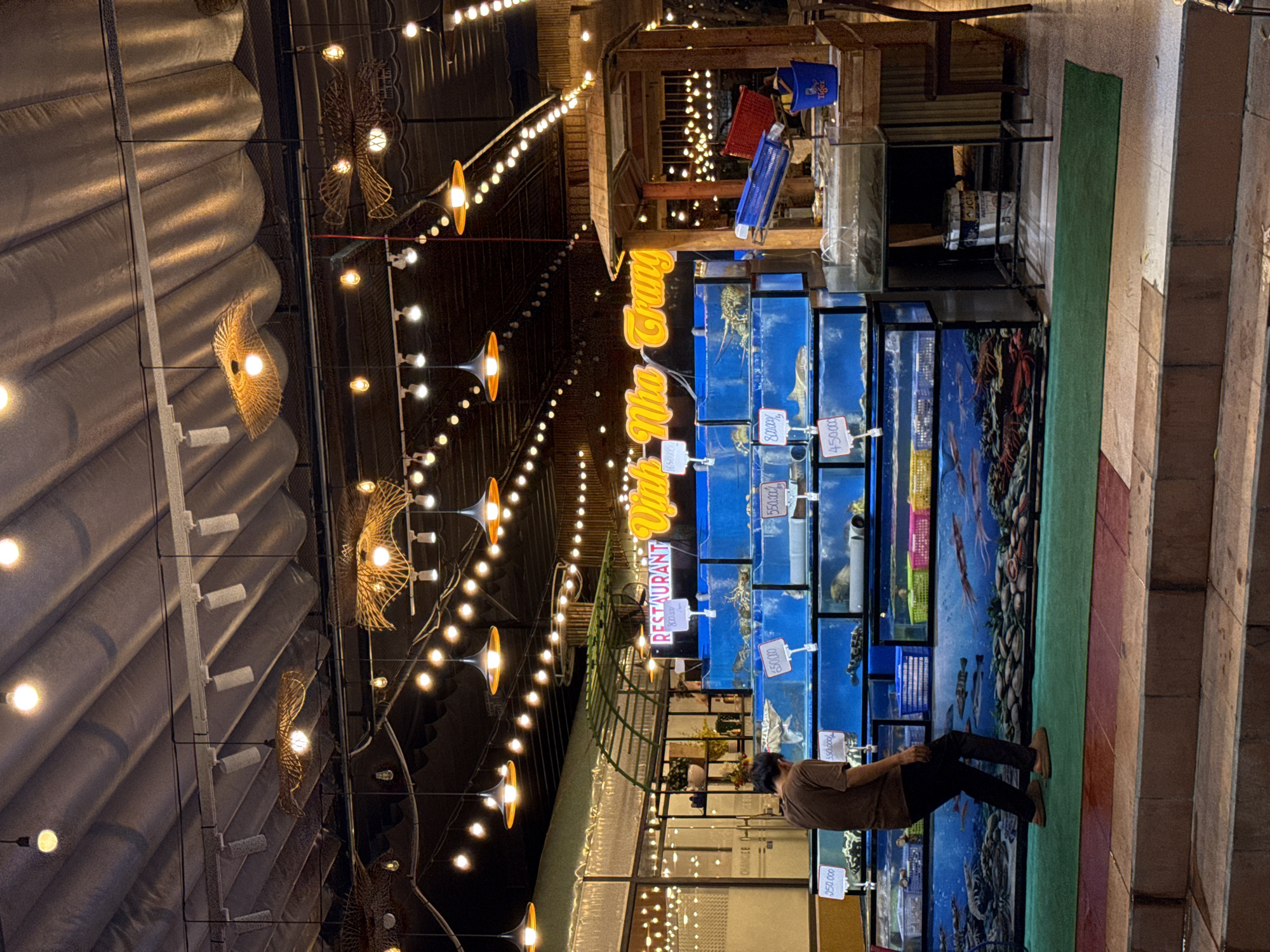 The image depicts a seafood restaurant in Nha Trang, Vietnam. A man in a brown t-shirt and black pants is standing to the left, looking at a display of live seafood in tanks. The tanks are filled with various types of marine life, including lobsters and fish, and are labeled with prices. The restaurant is decorated with hanging lights and woven lampshades. In the background, a neon sign reads "RESTAURANT Vinh Nha Trang". The overall scene is dimly lit, with the lights of the restaurant and the surrounding city creating a warm ambiance.