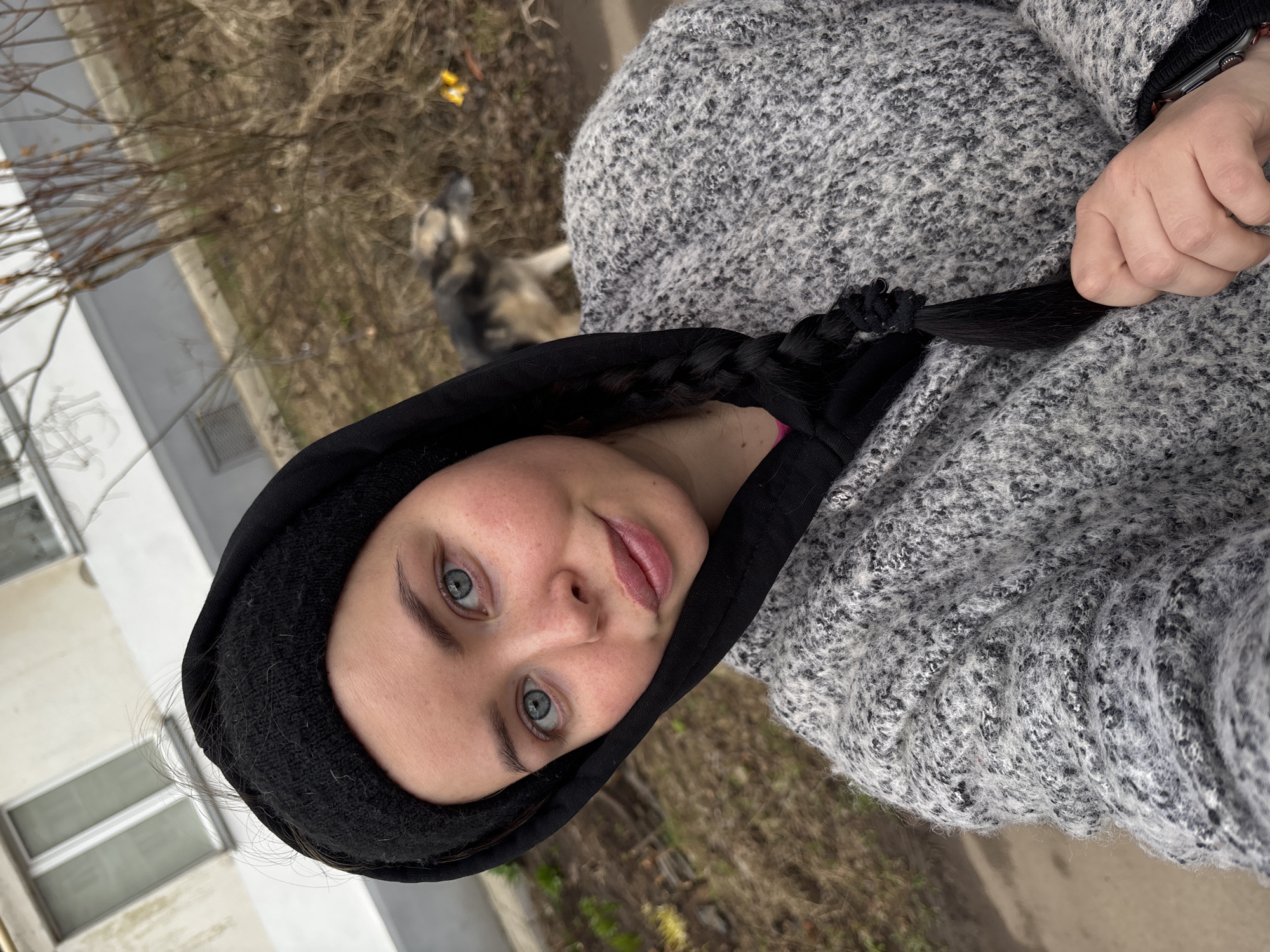 A woman wearing a black hooded beanie and gray sweater is centered in the image. She is turned slightly, looking directly at the camera with a subtle smile. The background includes bare trees, a patch of grass, and a corner of a building painted white. In the distance is what appears to be a dog. There are no signs of activity. Given the information and based on architectural styles, the scene is not likely to be in Izhevsk, Russia. There is no evidence to support this. The location is undetermined based on this photo.