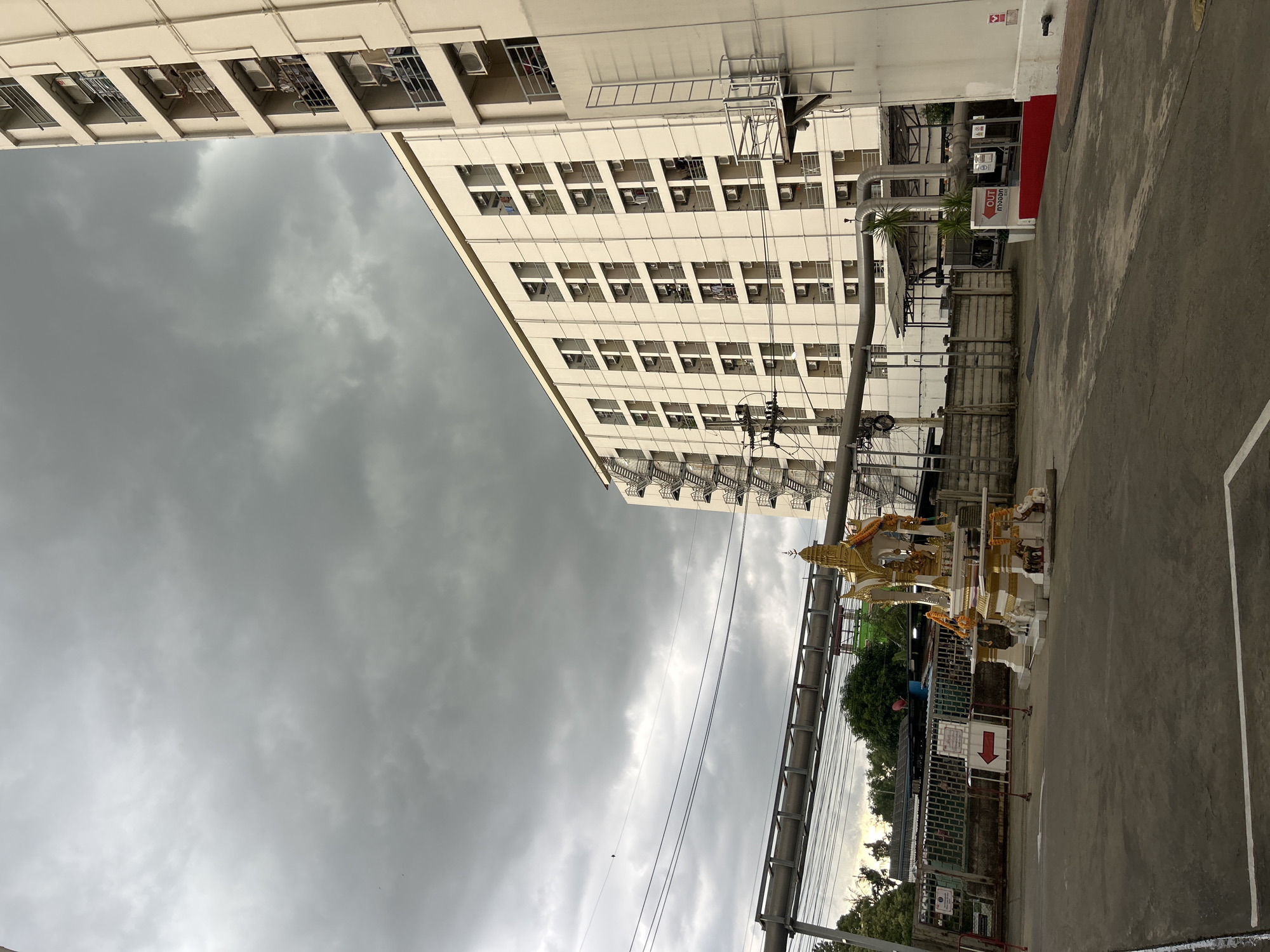 The image captures a scene on a cloudy day, likely in the city of Khlong Luang Town Municipality, Thailand. In the foreground, a paved area features a small, ornate shrine with a tiered, golden roof, decorated with garlands and supported by elephant figurines. A white and gold pedestal holds statues. To the left of the shrine, a red and white directional sign points to the left. Behind the shrine and sign, a concrete fence runs horizontally across the scene, with various structures and greenery visible beyond it. Dominating the background is a large, multi-story building with a cream-colored facade, numerous windows, and balconies. External staircases are visible on the left and right sides of the building. A network of pipes runs horizontally across the midground, supported by structures. Power lines and poles are also present. The overall impression is one of an urban or semi-industrial area, with a mix of residential buildings, infrastructure, and a cultural shrine.