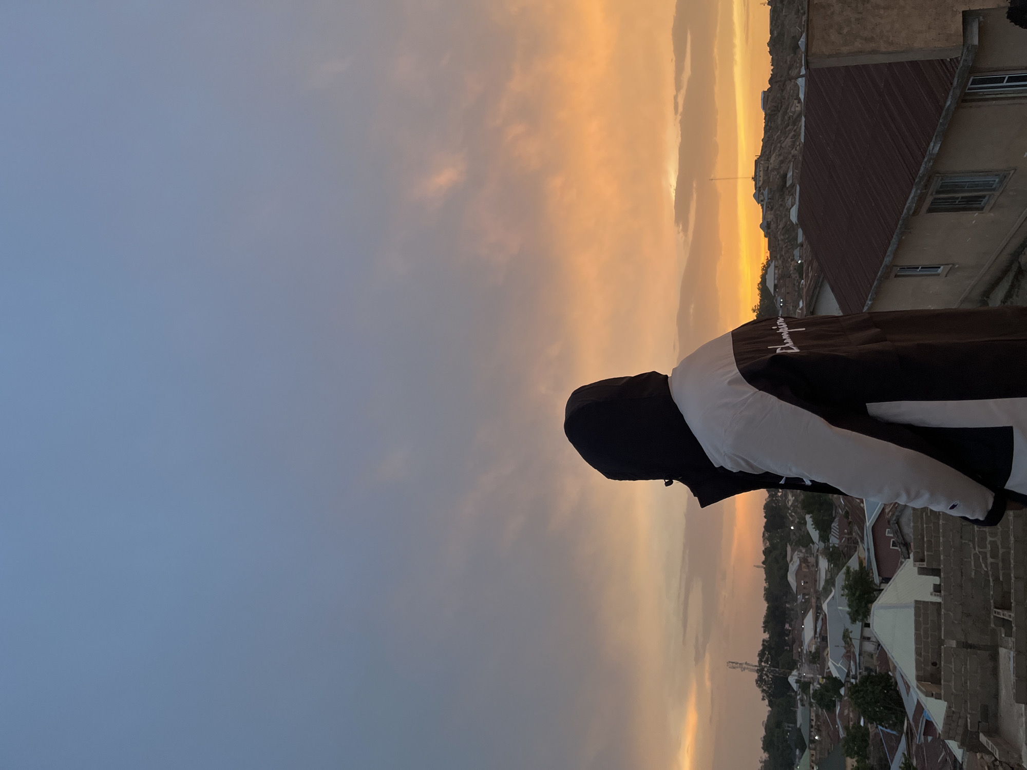 A vertical photograph depicts a person looking towards a city landscape during sunset. The person is wearing a black hooded sweatshirt, with the word "Champion" printed on the sleeve, and white accents on the sleeves. The city is Jos, Nigeria. The sky transitions from a light blue to an orange hue closer to the horizon. Buildings with brown rooftops are seen in the distance.