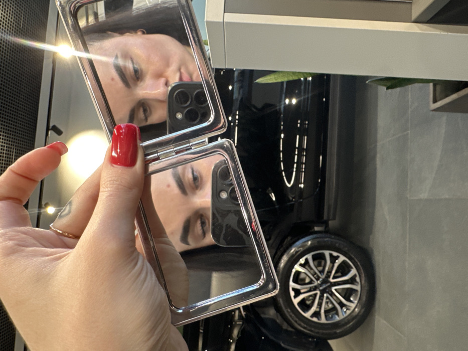 A person's hand, displaying bright red manicured fingernails and a ring on the ring finger, is holding a silver-framed compact mirror. The mirror reflects the upper portion of a female face, specifically the eyes, dark eyebrows, and part of the nose and cheeks, along with the back of a smartphone featuring a multi-lens camera module. The person in the reflection appears to be looking downwards. In the background, a glossy black car is visible, featuring multi-spoke silver and black rims on its front wheel. The environment is an indoor space with a dark gray tiled floor. A light-colored vertical pillar and a partially visible potted plant are present on the right side. Reflections of overhead grid lighting are discernible on the car's surface and within the mirror, suggesting a commercial setting such as a car showroom. The specific location within Saint Petersburg, Russia, cannot be determined from the visual information provided in the image.