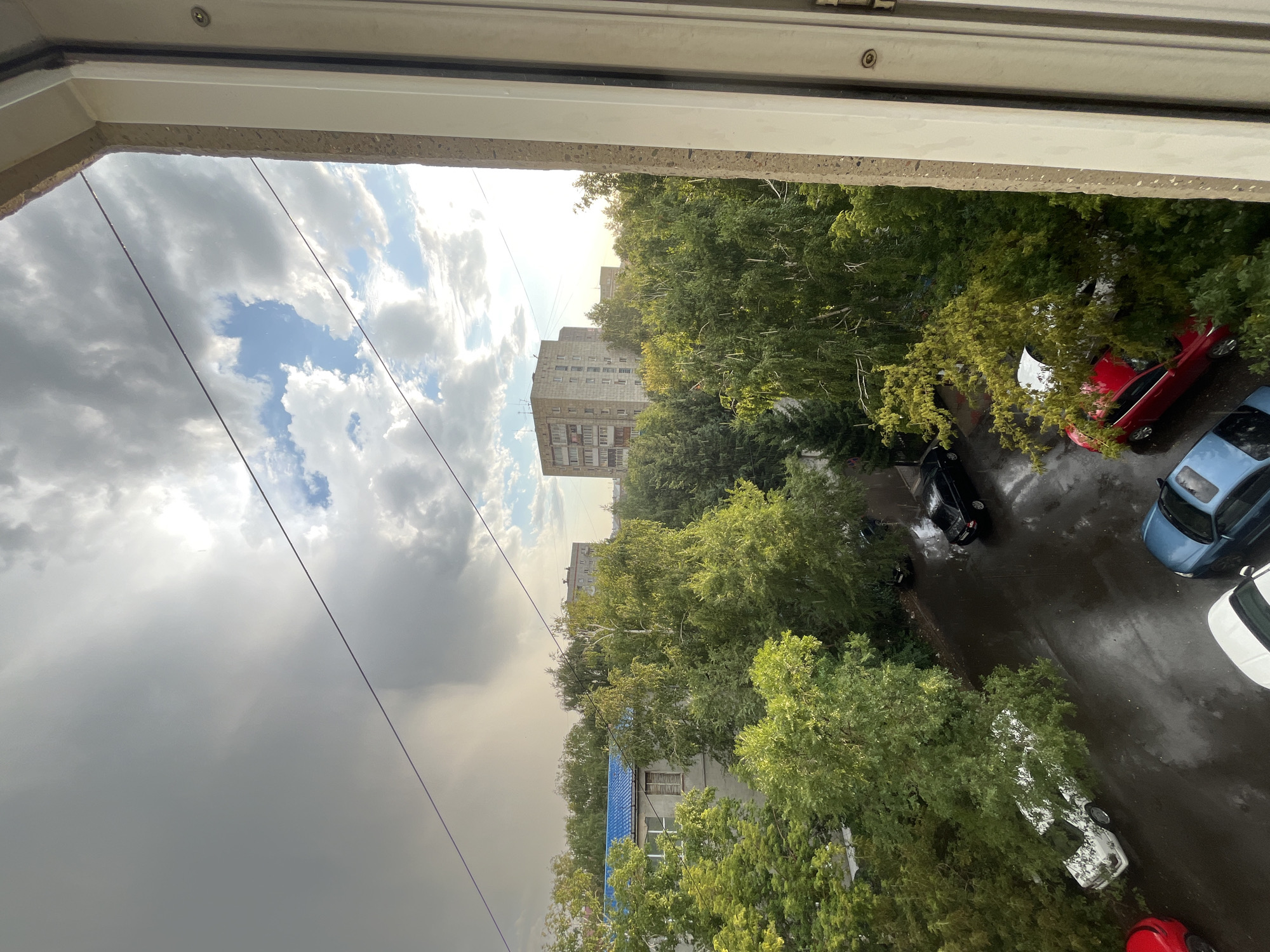 The image captures a view from a window overlooking a city street in Oskemen, Kazakhstan, during a rain shower. The sky is filled with dramatic clouds, with a patch of sunlight breaking through on the right. A heavy rain curtain is visible in the distance on the left. Trees with lush green foliage line the street, and several parked cars are scattered on the wet asphalt. The buildings in the background appear to be residential apartment blocks. The foreground shows a window frame on the right side of the image. No people are visible in the scene.
