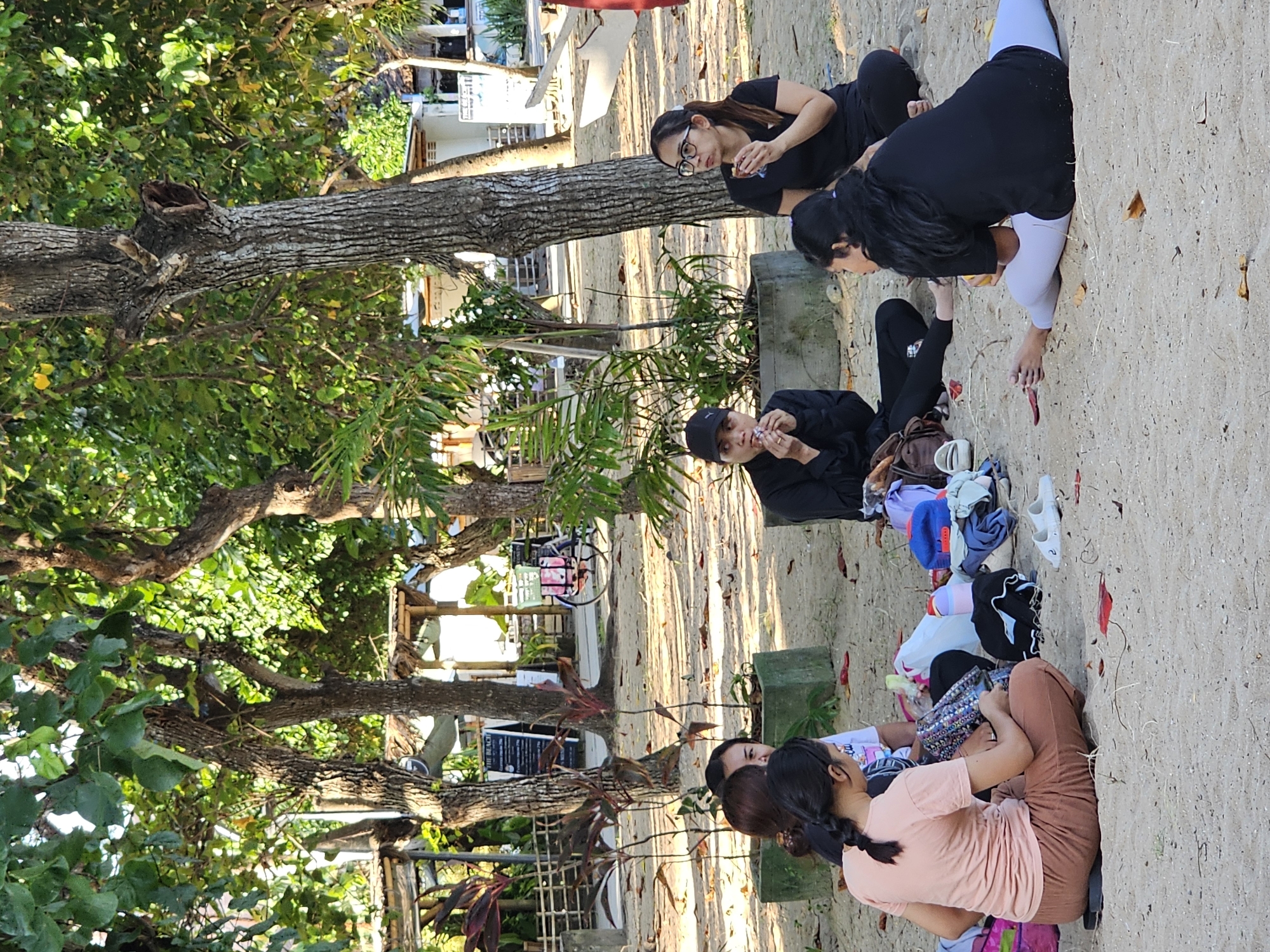 A daytime eye-level shot captures a group of people sitting on the beach in Denpasar, Indonesia. The group, comprising five females, is situated on the sandy ground, in the center. One person is wearing glasses and a black shirt, one is in a light pink shirt and brown pants, one is wearing black pants and a black sweatshirt, and another has a black shirt and white pants. The fifth person is partially visible. They are surrounded by a collection of personal items including bags, clothes, and shoes scattered around them on the sand. The background features a lush green tree, a row of buildings, and beach vegetation. The composition emphasizes the social interaction among the individuals within the coastal environment.