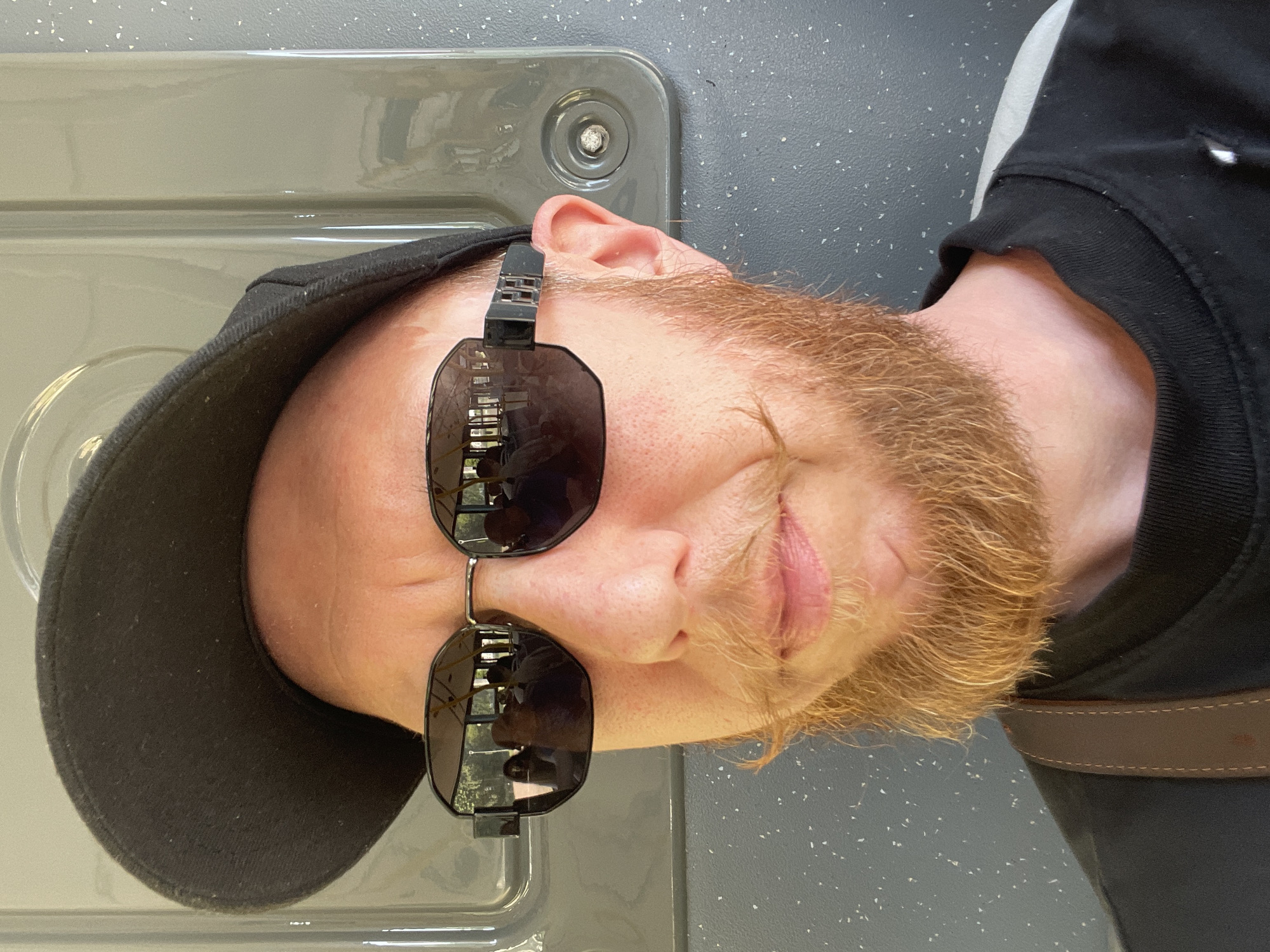 A person with reddish-blonde hair and beard, wearing a black baseball cap and black sunglasses, is pictured in a close-up, sideways shot. The sunglasses are black with a thick frame, reflecting a blurred image of trees and possibly buildings. The person is wearing a black hoodie or sweatshirt, and a brown leather strap, possibly from a bag, is visible across their shoulder. The background appears to be an indoor setting with a textured, speckled grey surface and a smooth, light grey panel with a circular fixture. The lighting suggests it could be daytime, but the indoor setting makes it difficult to determine. The image is a selfie, with the camera angled from above. The person's expression is difficult to ascertain due to the sunglasses and sideways orientation. No visible text is present in the image.