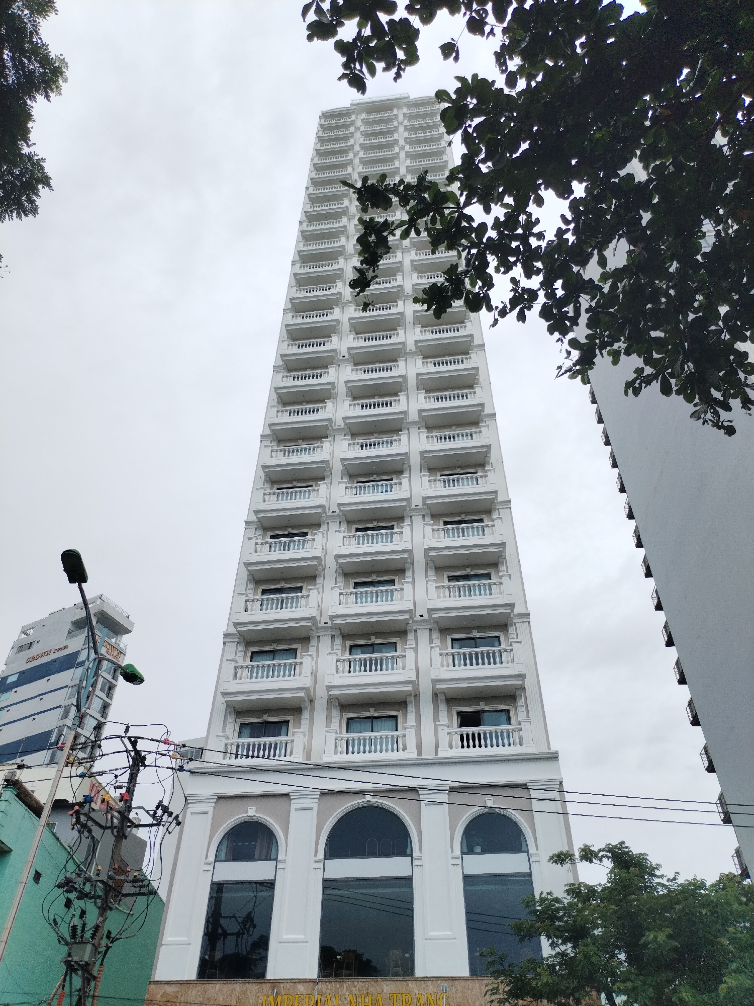 The image features a tall, white building with many balconies in Nha Trang Ward, Vietnam. The building has a facade of large arched windows on the ground level and is flanked by other buildings and trees. Overhead power lines and a street lamp are visible in the foreground. The sky is overcast. The name "IMPERIAL NHA TRANG" is visible above the building's entrance.